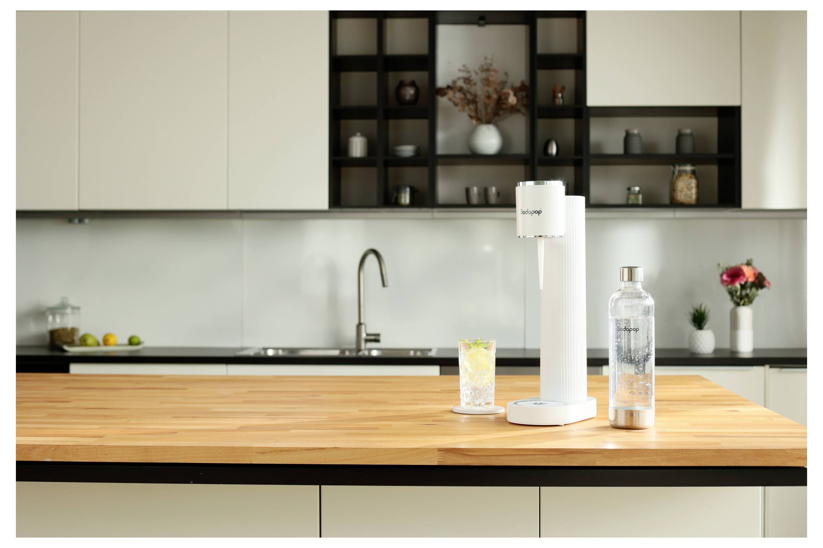 Eine weiße Küchenarbeitsplatte mit einem Getränkecarbonator, einem gefüllten Glas und einer Flasche im Fokus. Moderne Küchenschränke und Dekoration im Hintergrund.