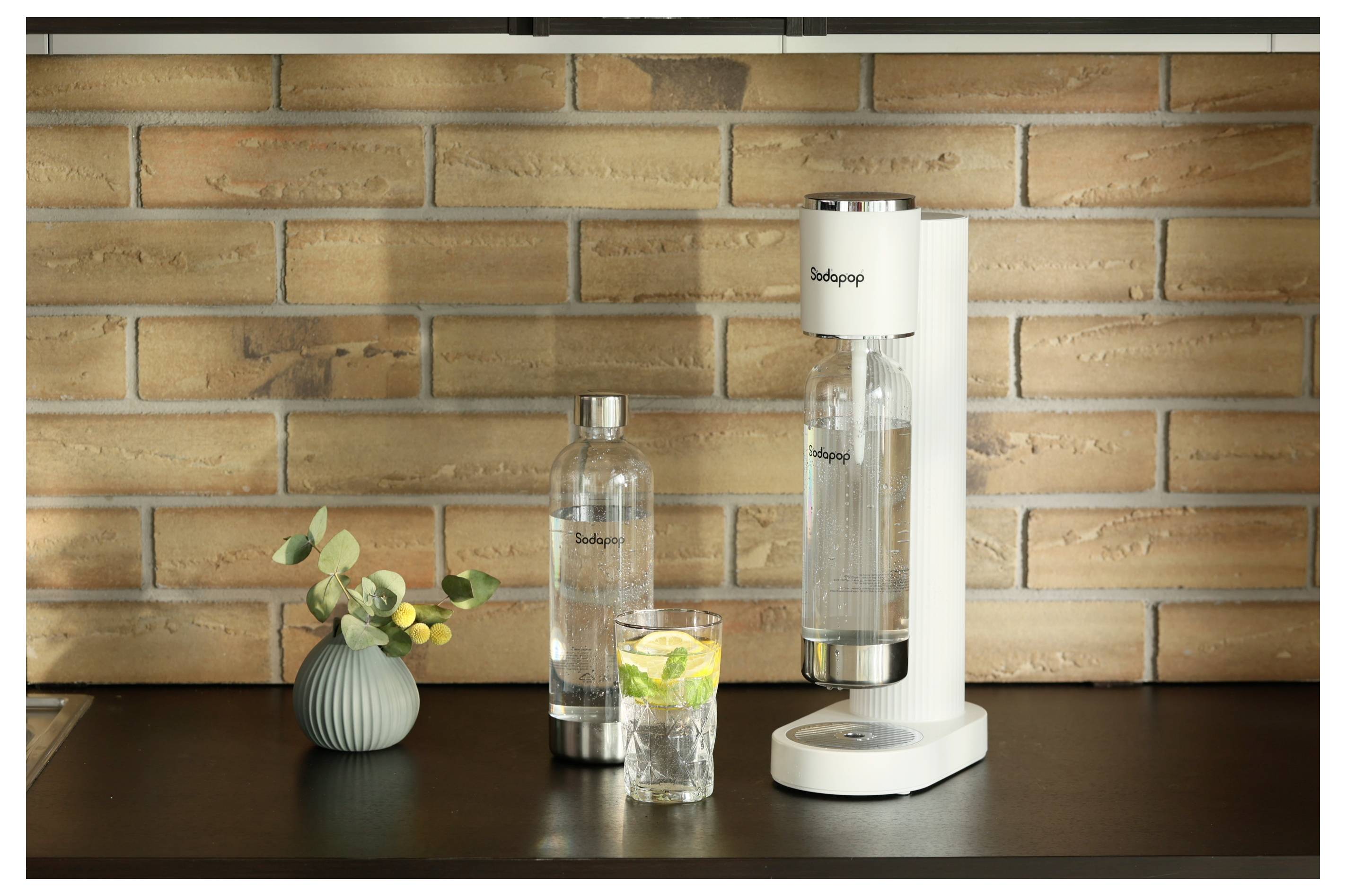 Ein weißer Sodahersteller steht auf einer Küchenarbeitsplatte vor einer Ziegelwand, zusammen mit zwei Flaschen und einem Glas mit Sprudelwasser und Limette.