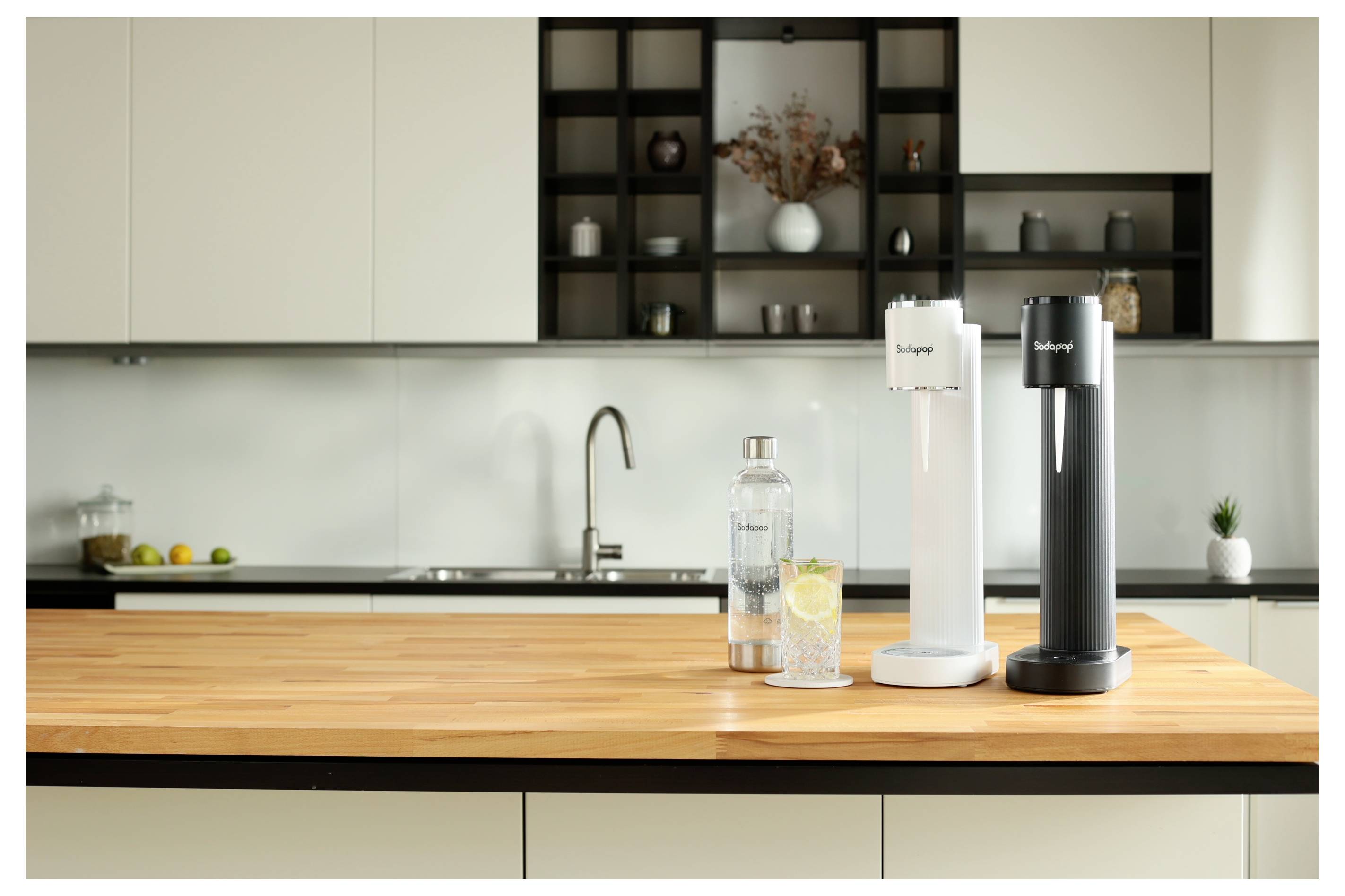 Eine moderne Küche mit zwei Sodamaschinen auf einer Holzarbeitsfläche, begleitet von einer Glasflasche und einem Wasserglas mit einer Zitronenscheibe.