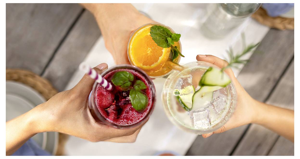 Drei Hände erheben farbenfrohe Getränke in einer Feiergestik. Die Getränke umfassen ein rotes Beerengetränk, einen Cocktail mit Orangenscheibe und ein Gurken-infundiertes Getränk.