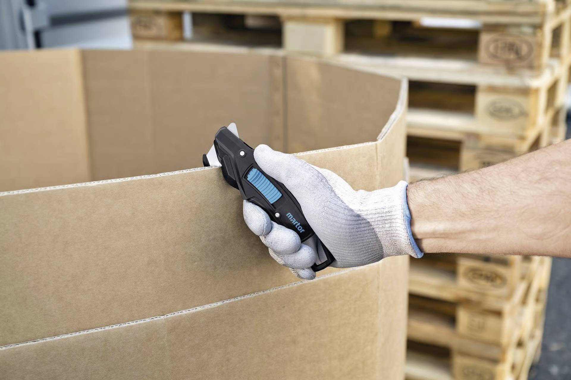 Eine Person mit Handschuh verwendet ein Utility-Messer, um einen großen Karton zu schneiden. Im Hintergrund sind Holzpaletten gestapelt.