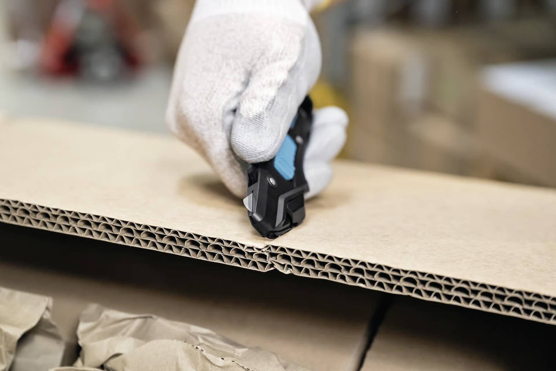 Eine Handschuh tragende Hand verwendet ein Universalmesser, um einen Karton zu öffnen und braunes Packpapier im Inneren in einem Lagerhausumfeld freizulegen.