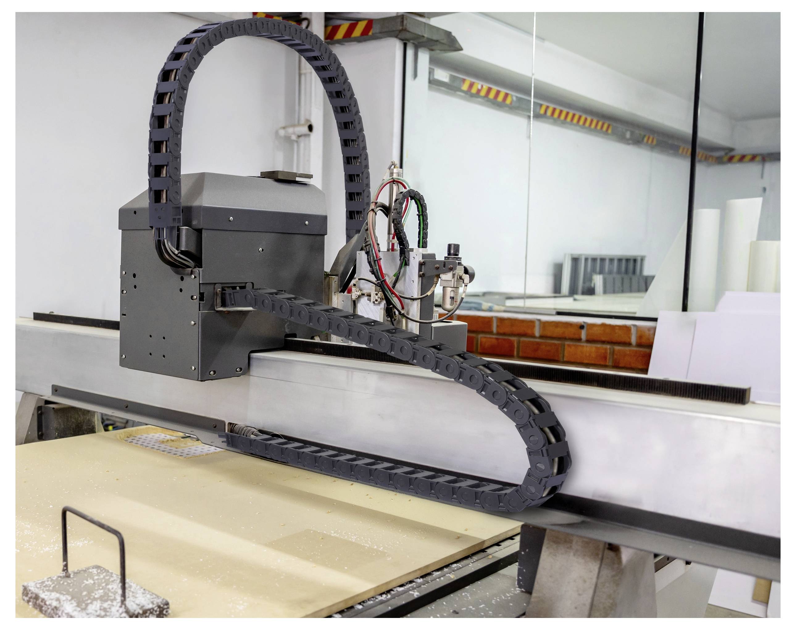Eine große CNC-Maschine, die ein Holzpaneel schneidet. Die Maschine hat eine schwarze, flexible Kabelstrecke und befindet sich in einer Werkstattumgebung mit sichtbaren Werkzeugen.
