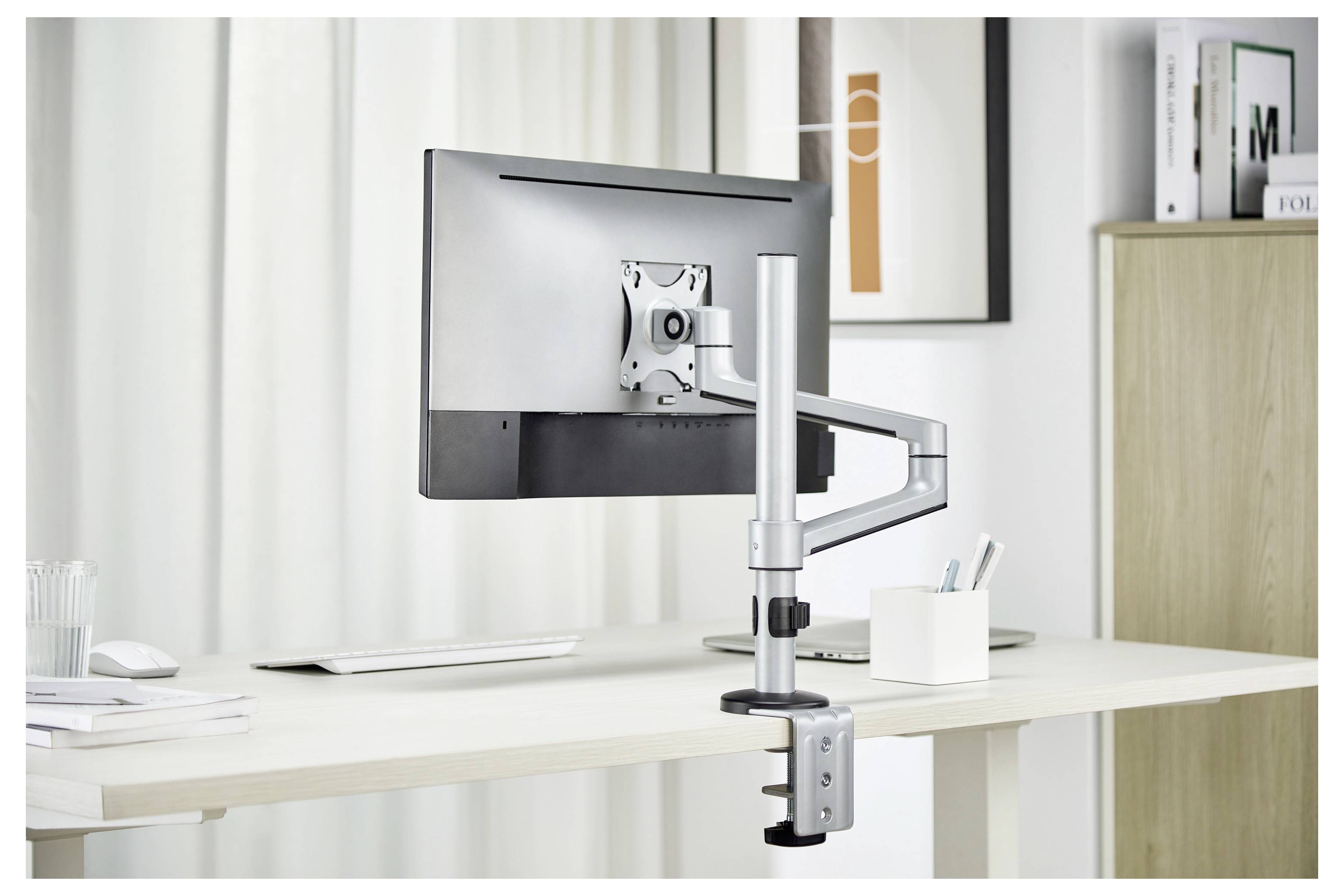 Ein verstellbarer Monitorarm, der einen Computerbildschirm über einem weißen Schreibtisch hält, mit einem Regal und gerahmtem Kunstwerk im Hintergrund sichtbar.
