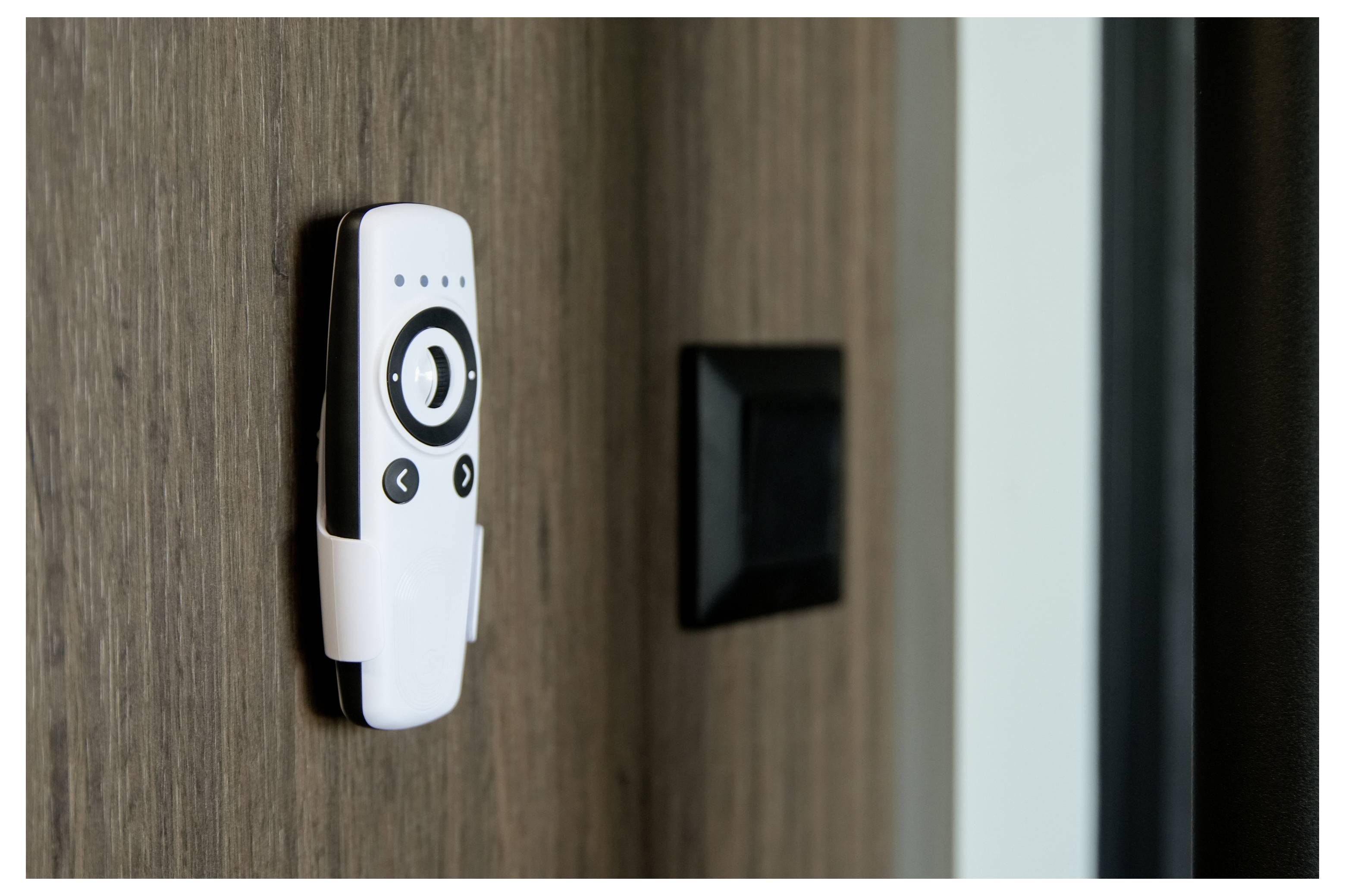 Eine Fernbedienung mit Tasten ist an einer Holzwand neben einem schwarzen Schalter angebracht, was auf die Steuerung von Haushaltsgeräten oder Beleuchtung hindeutet.