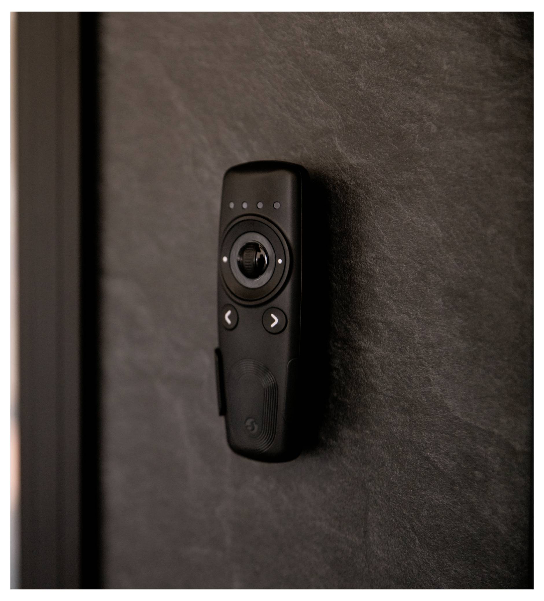 Eine schwarze Fernbedienung, die auf einer dunklen strukturierten Wand montiert ist und Tasten sowie ein zentrales kreisförmiges Steuerpad aufweist.