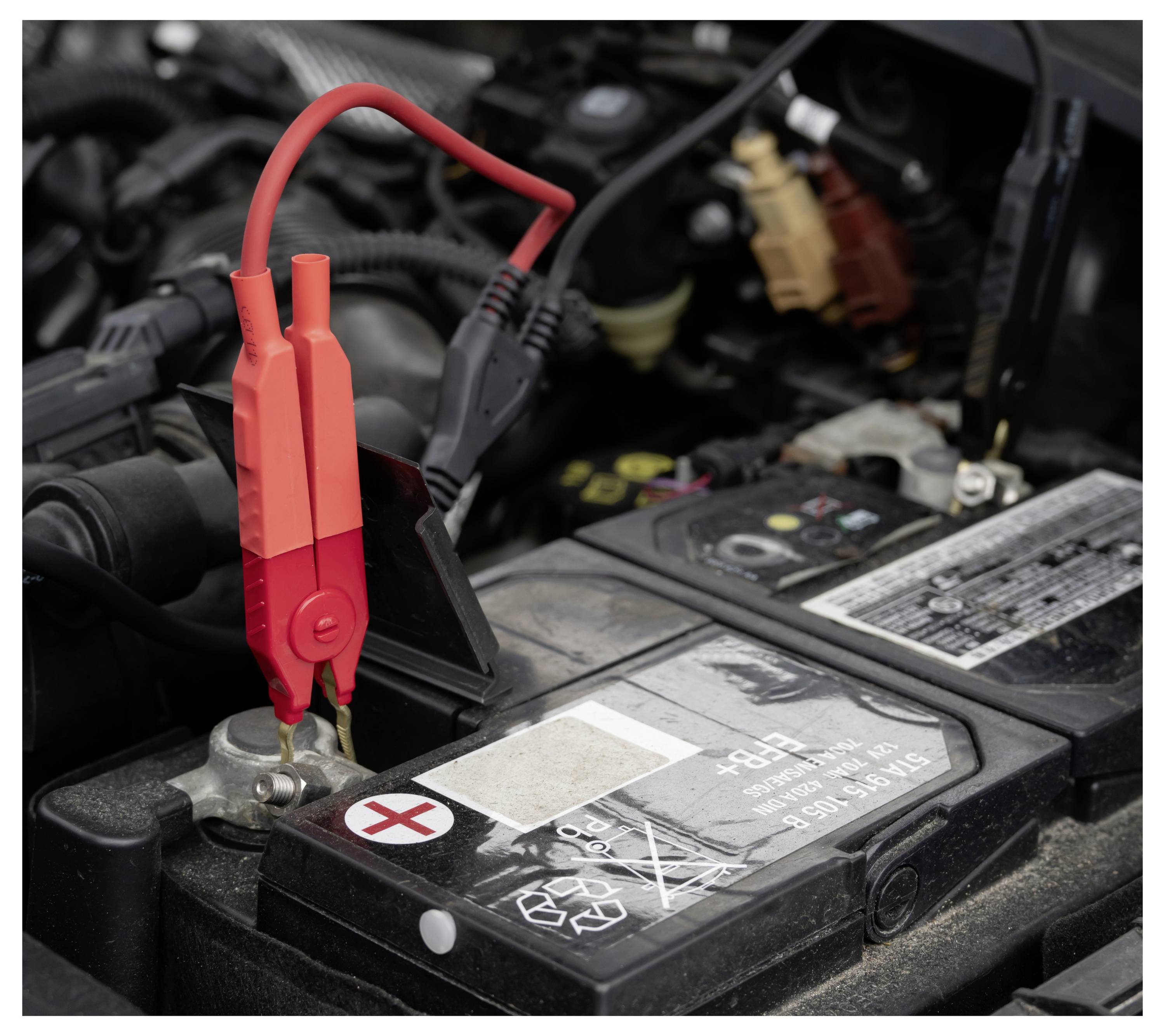 Rote Starthilfekabel an einer Autobatterie in einem Motorraum angeschlossen, was auf Starthilfe oder das Aufladen der Batterie hindeutet.