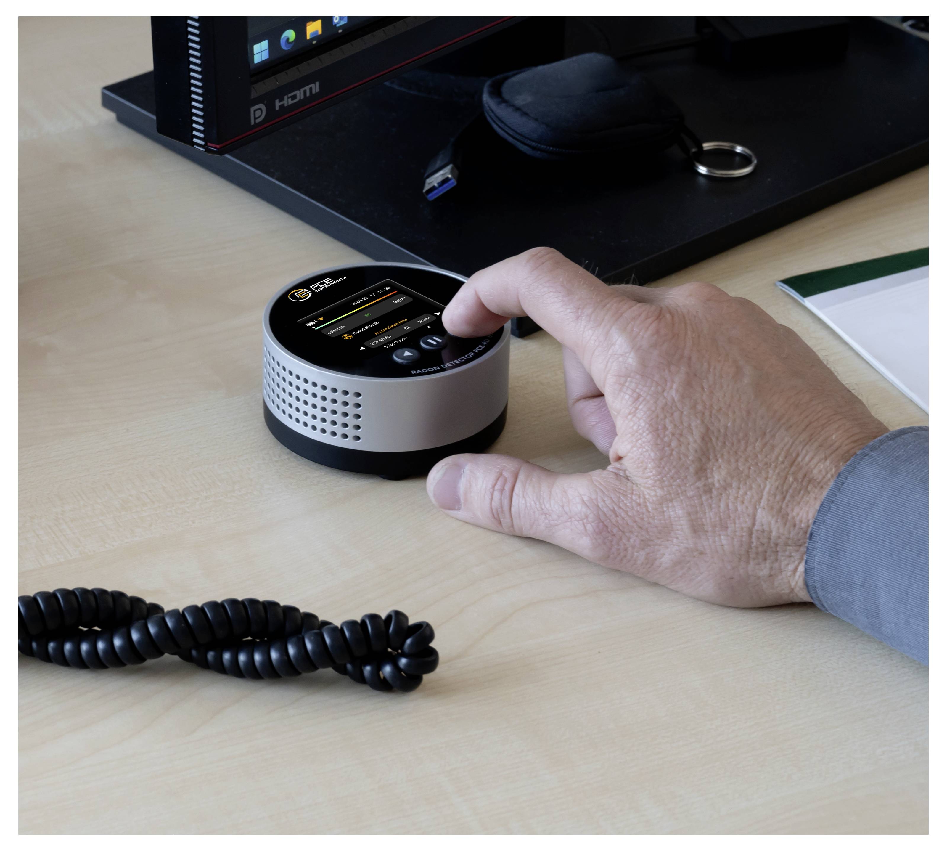 Eine Hand drückt ein kleines rundes elektronisches Gerät auf einem Schreibtisch, mit einem Computermonitor und Zubehör im Hintergrund.