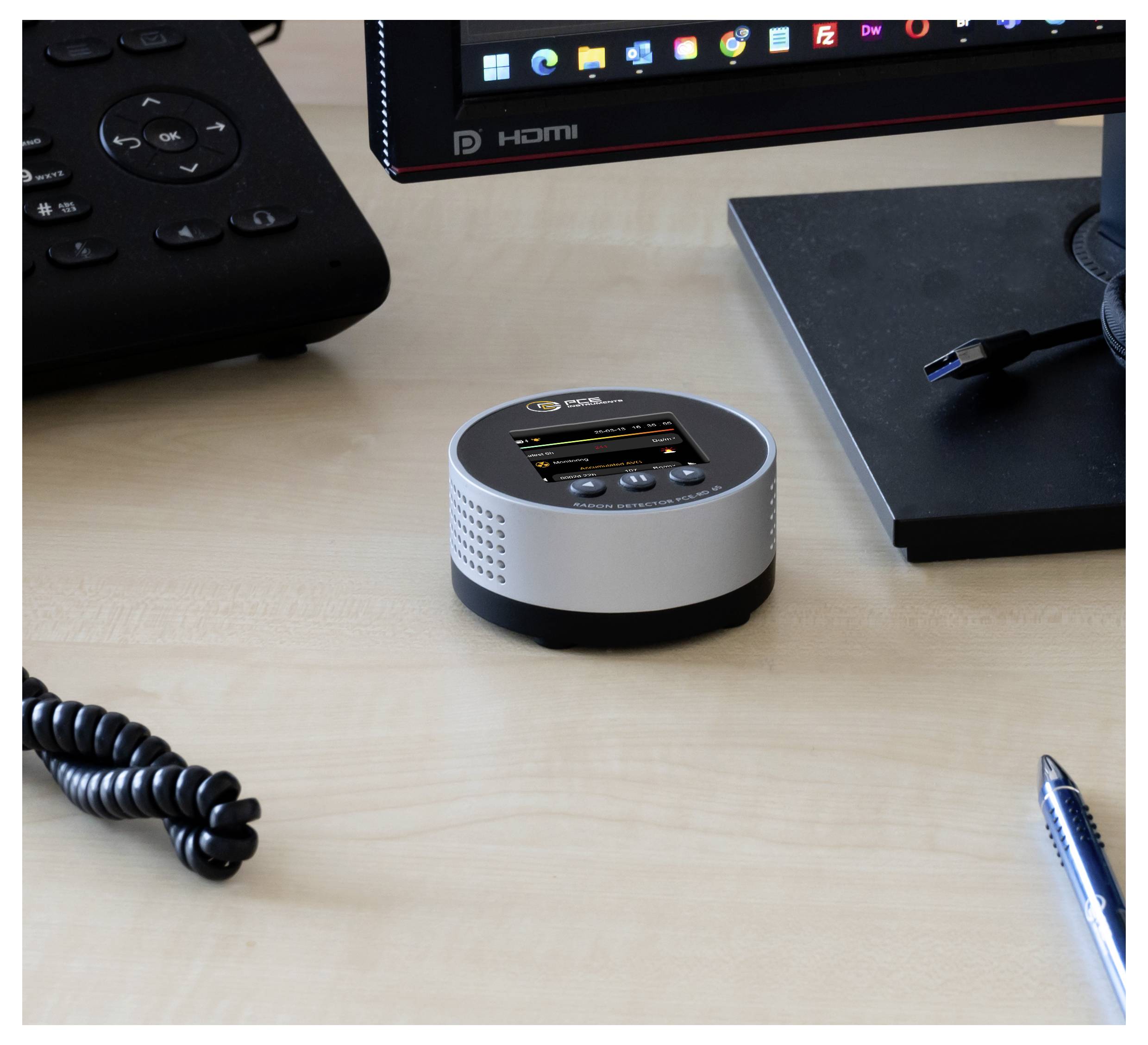 Kleines rundes silberfarbenes Tischgerät mit winzigem Display und Tasten auf einem Schreibtisch neben einem Monitor, einer Fernbedienung, einem Stift und einem Kabel.