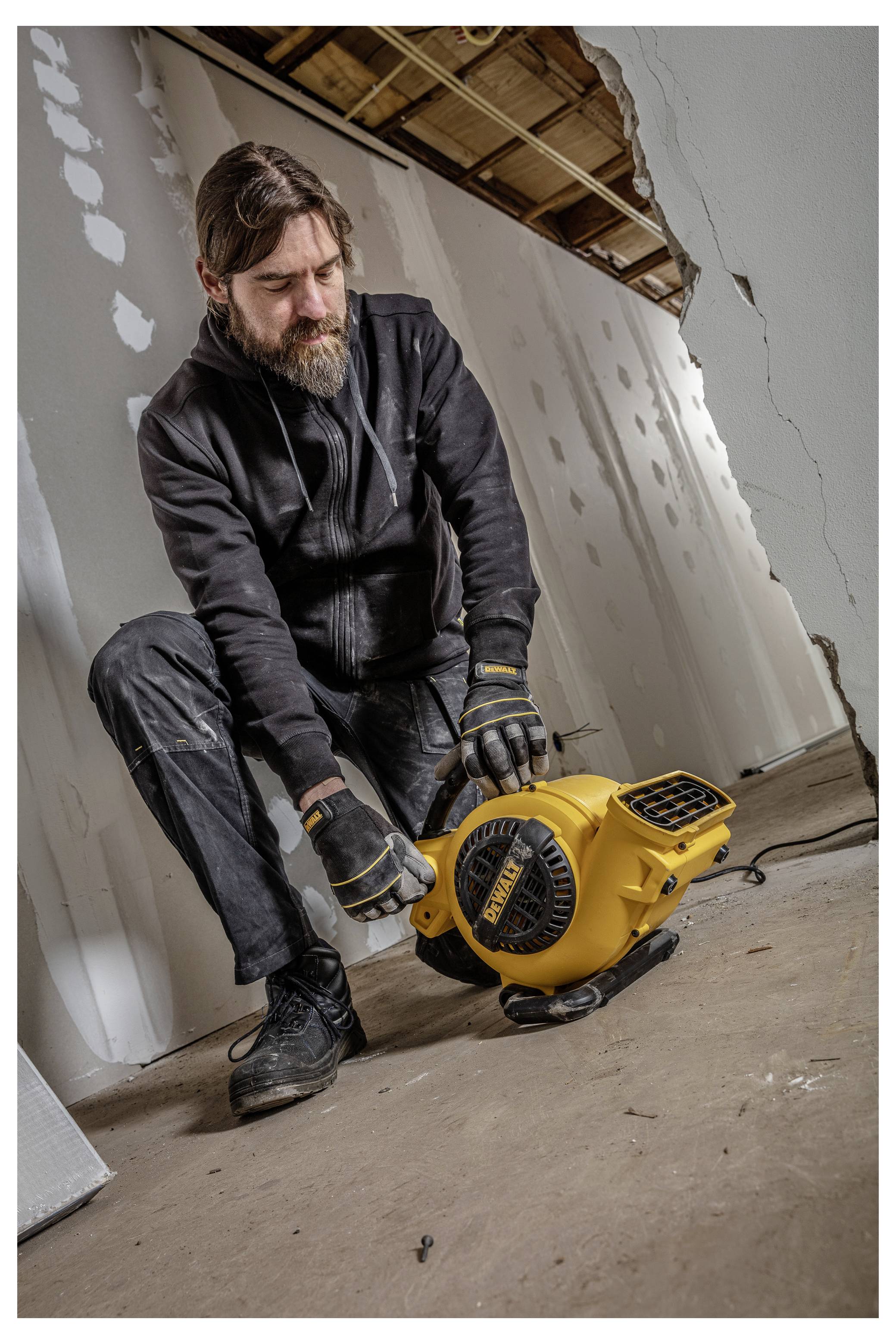 Ein bärtiger Mann in Arbeitskleidung stellt einen gelben Bodenventilator oder Lüfter in einem Raum unter Renovierung mit freiliegenden Deckenbalken und unvollendeten Wänden ein.