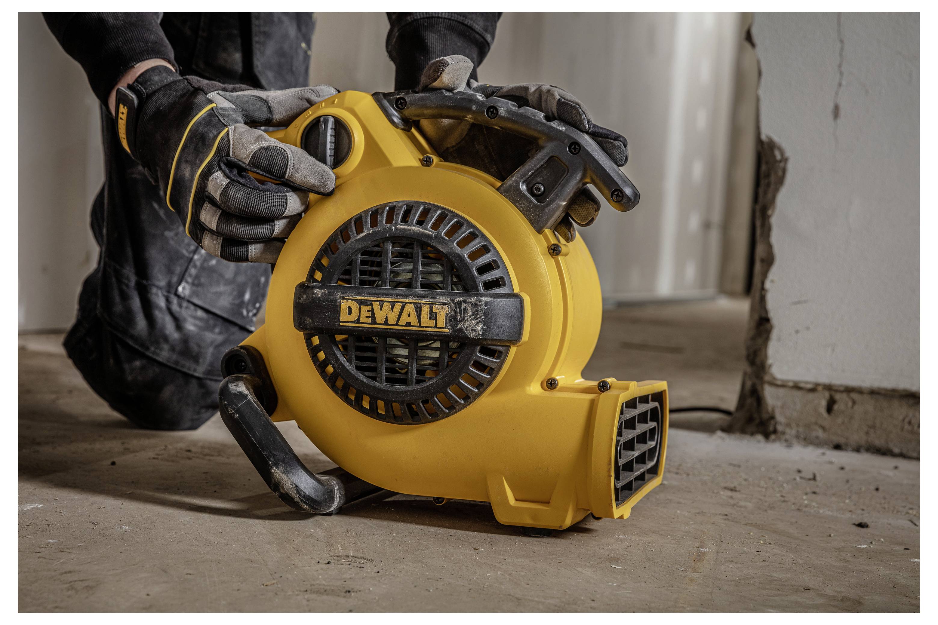 Handschuhgetragener Arbeiter, der ein gelbes tragbares DEWALT-Gebläse/Ventilator auf einem Baustellen-Fußboden bedient.