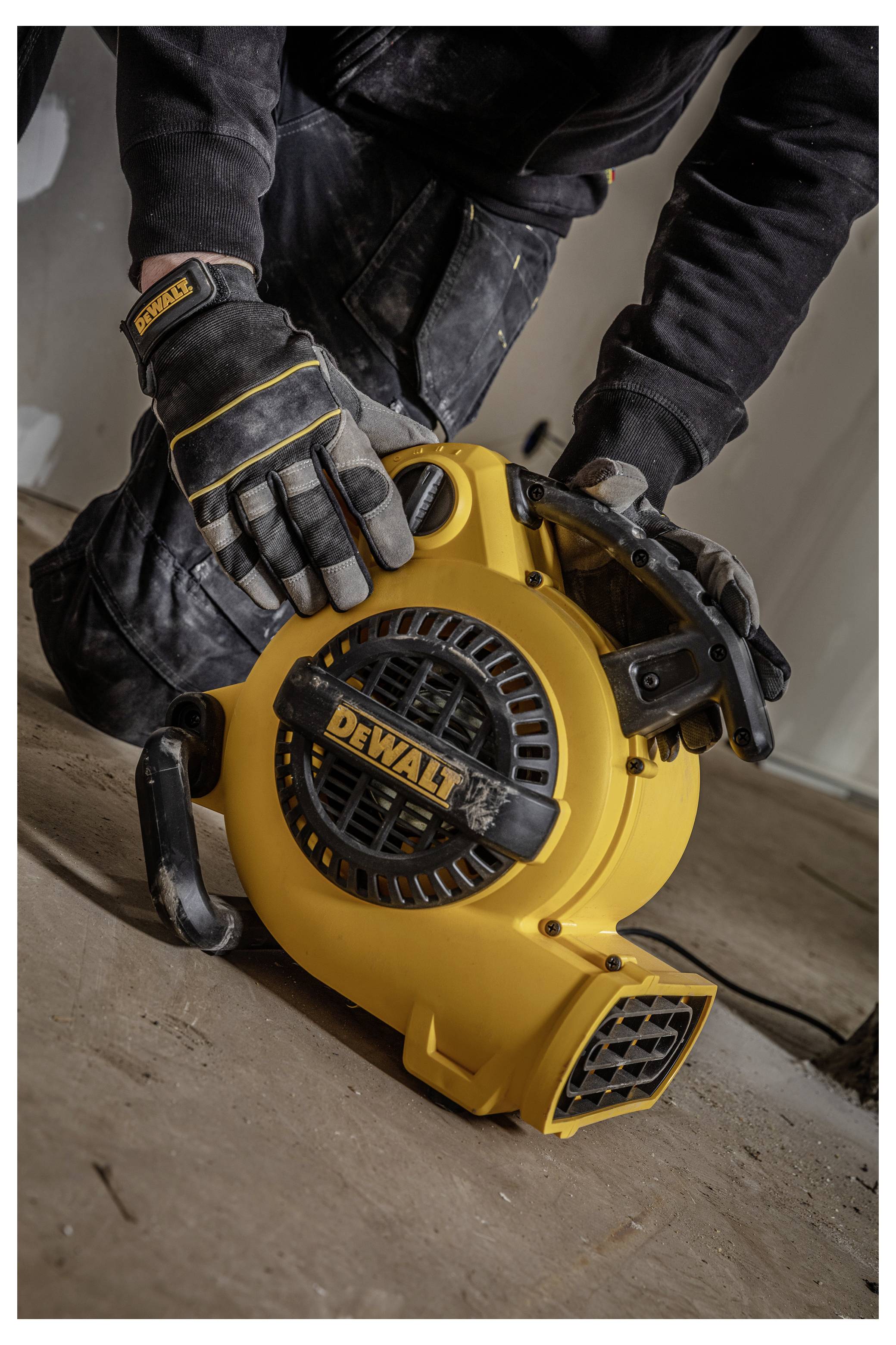 Handschuhtragende Arbeitskraft, die ein gelbes tragbares DEWALT-Gebläse auf einem staubigen Boden während Aufräumungs- oder Bauarbeiten bedient.