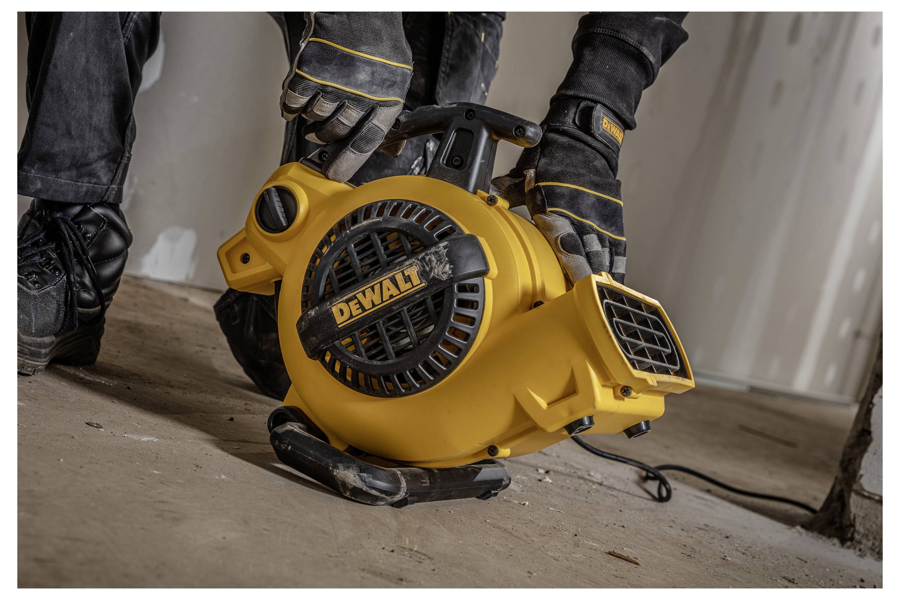 Ein Arbeiter mit Handschuhen hält ein gelbes tragbares DEWALT-Gebläse oder einen Ventilator auf einem staubigen Boden während einer Bau- oder Reinigungsarbeit.