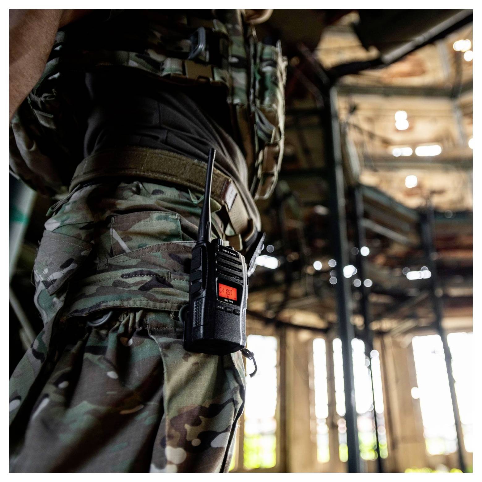 Eine Person in militärischer Tarnkleidung mit einem an ihrem Gürtel befestigten Funkgerät steht in einem Industriegebäude, durch dessen Fenster Sonnenlicht hereinströmt.