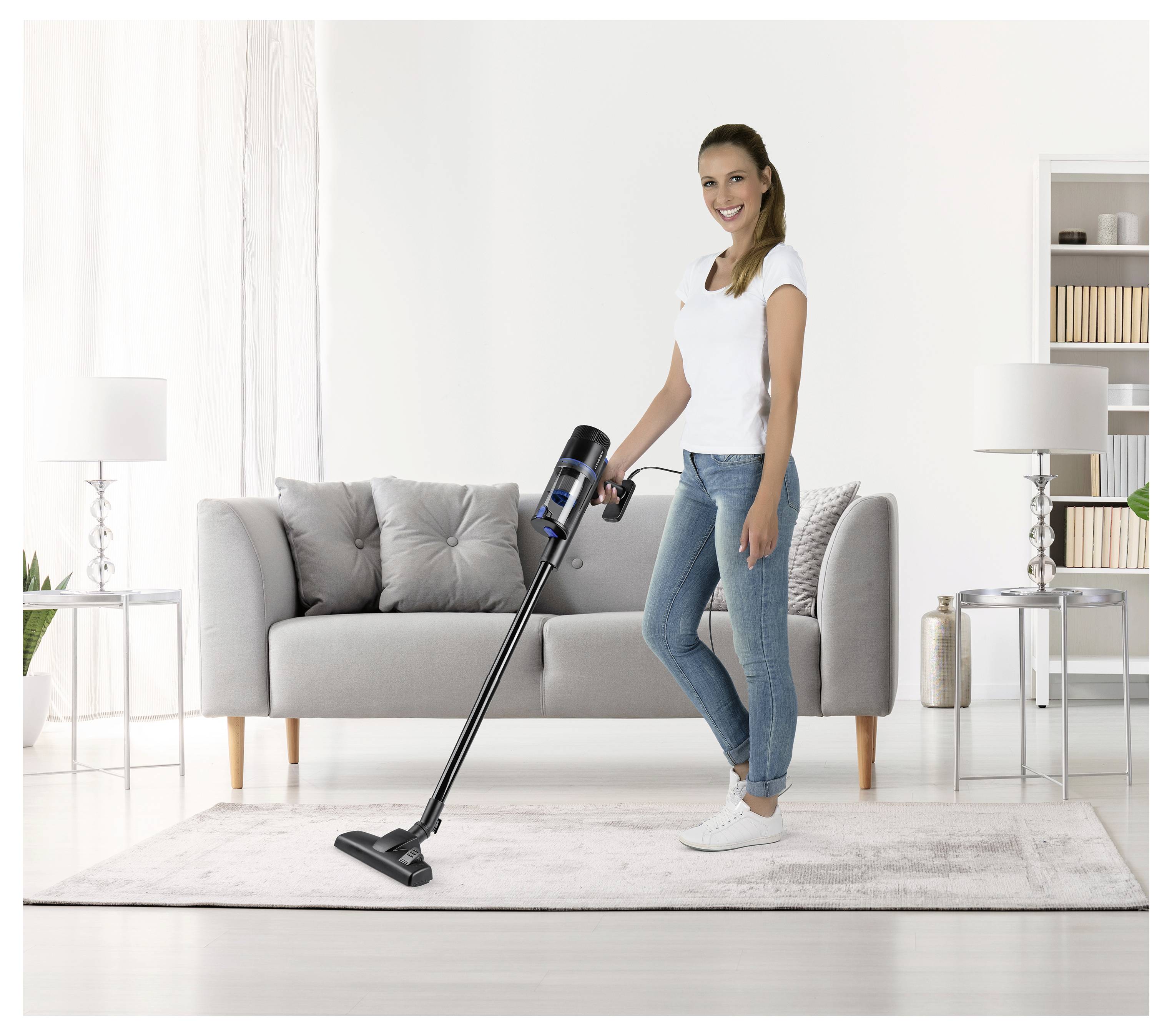 Eine Person saugt einen hellfarbigen Teppich in einem Wohnzimmer mit einem grauen Sofa, zwei Lampen und einem Bücherregal im Hintergrund ab.