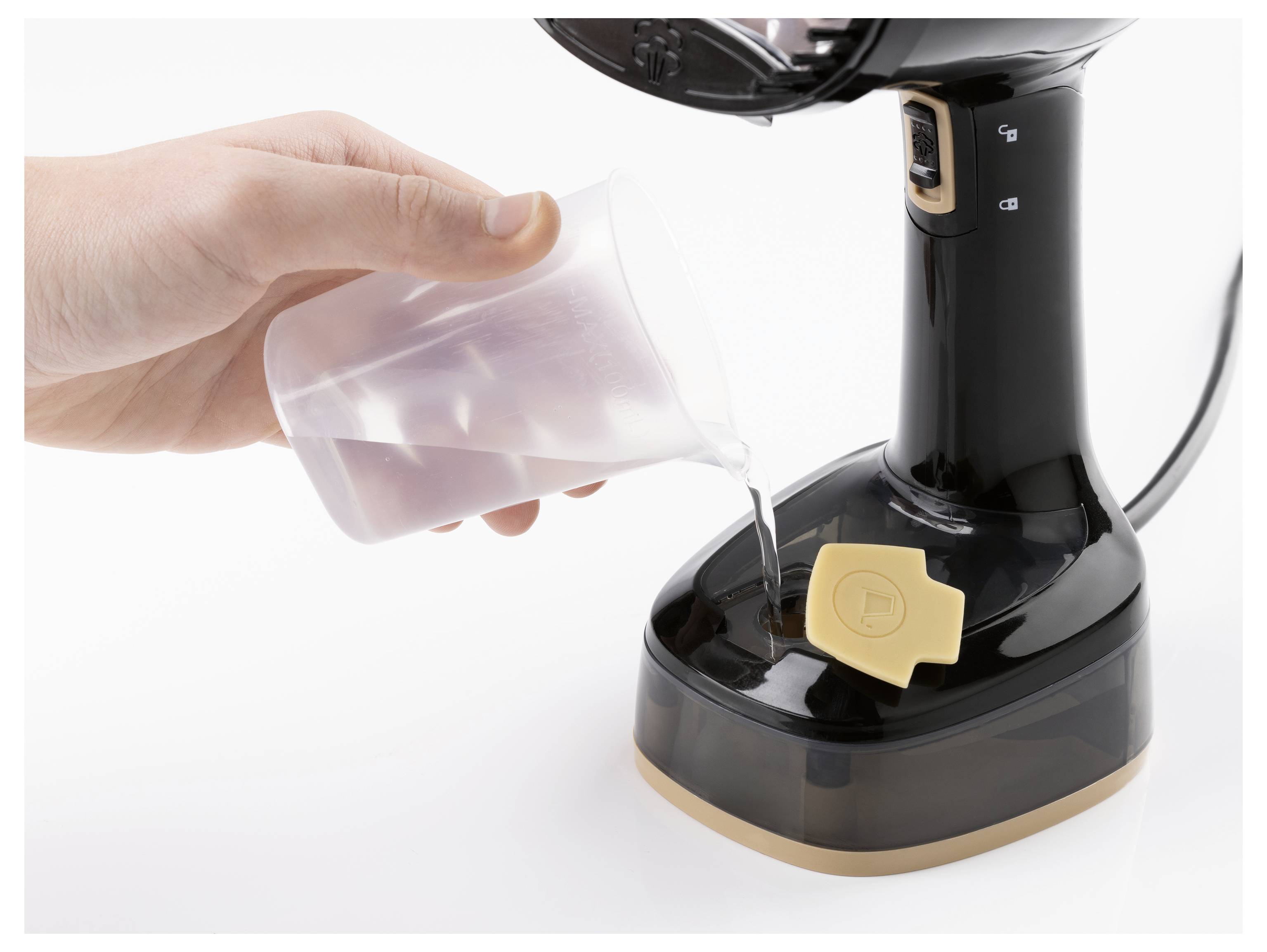 Eine Hand gießt Wasser aus einem Messbcher in den Tank eines schwarzen Dampfgeräts mit einem beigen Knopf, wahrscheinlich ein Kleiderdampfer.