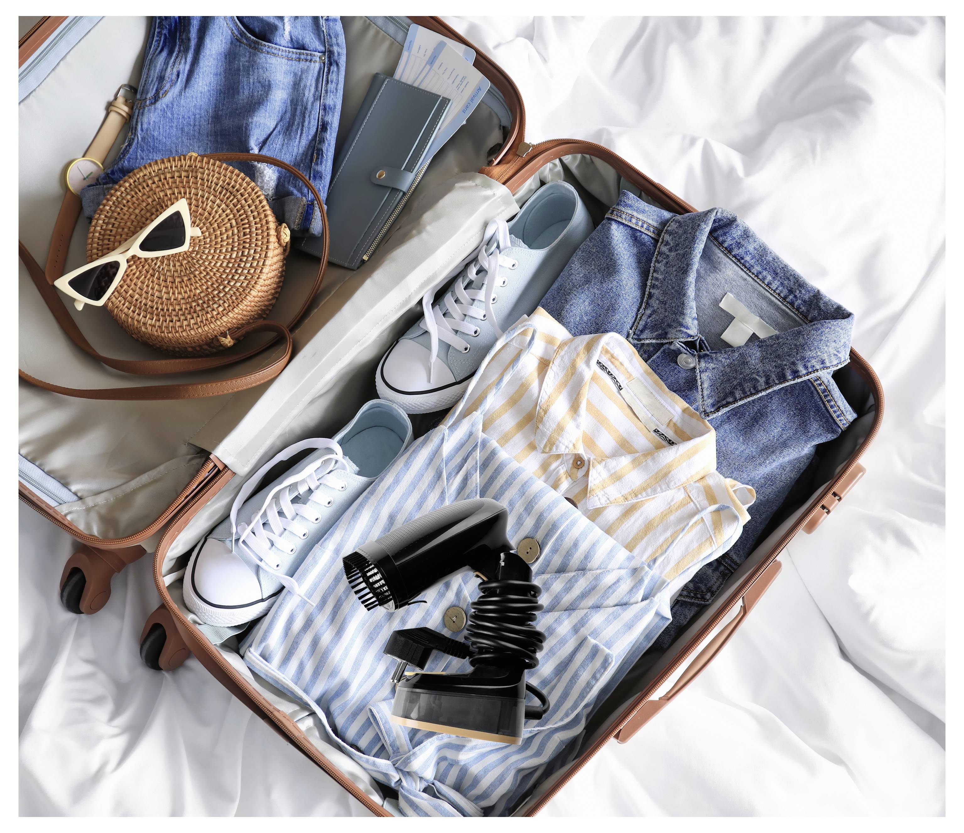 Offener Koffer auf dem Bett mit ordentlich gepackter Kleidung, Turnschuhen, einem Jeanshemd, gestreiften Tops, einem Föhn, einer runden geflochtenen Tasche und einer Sonnenbrille.