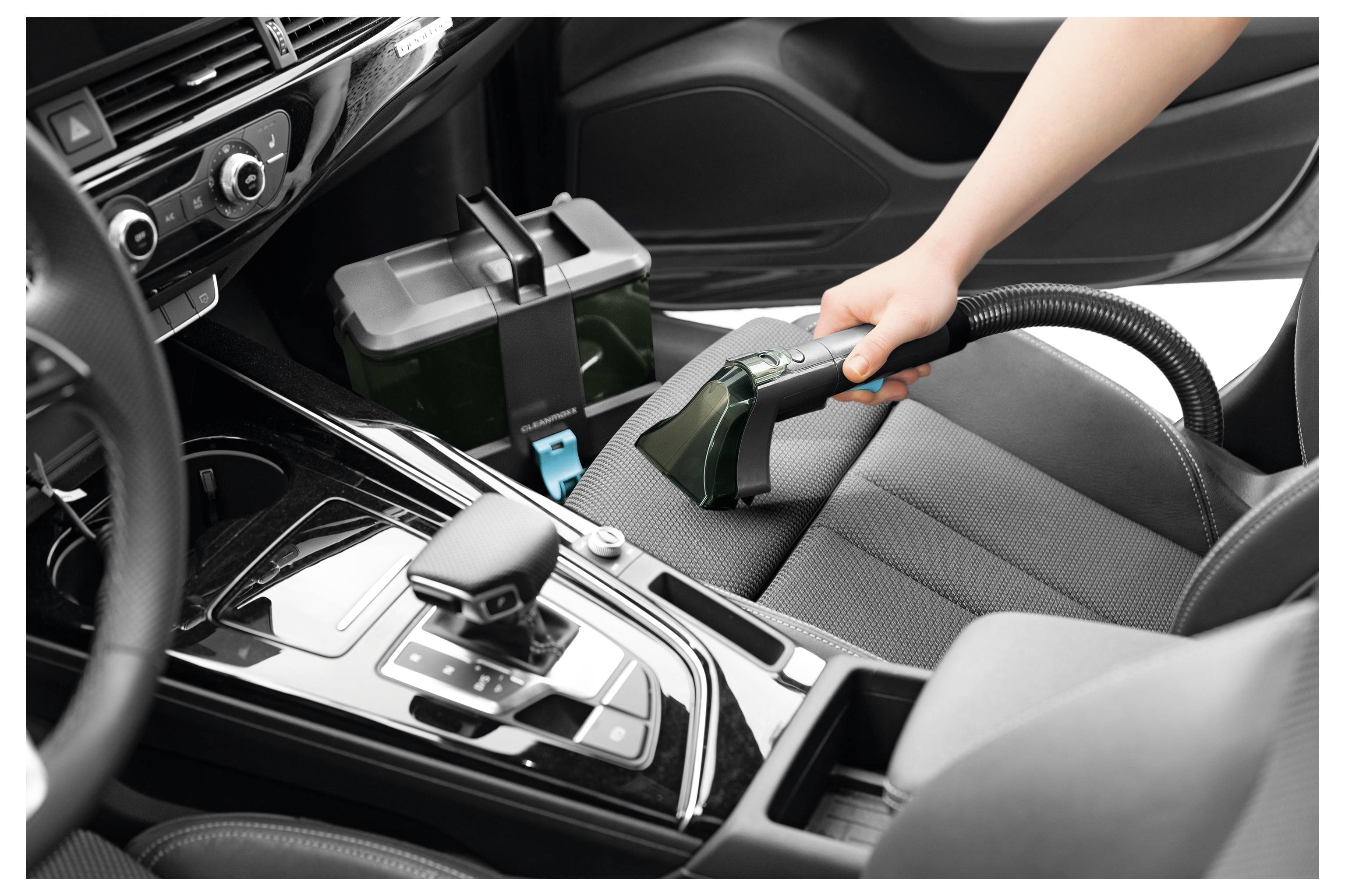 Autoinnenraum absaugen: Hand mit kleinem Staubsauger auf schwarzem Sitz, in der Nähe der Schaltkulisse und der Autobatterie.