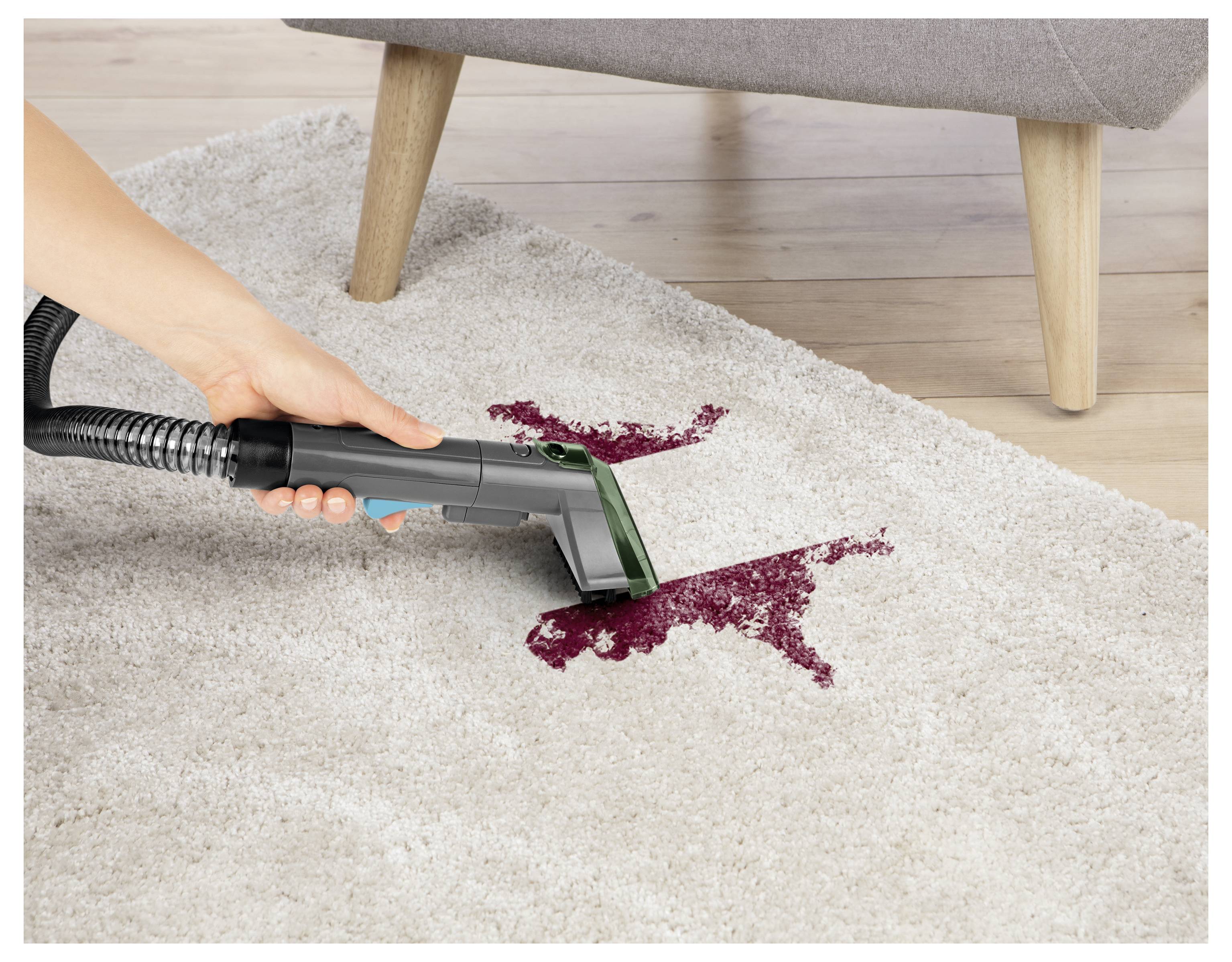 Ein Staubsauger mit langem Stiel reicht unter ein graues Sofa auf Holzbeinen. Der Fokus liegt auf der Reinigung von engen Räumen.