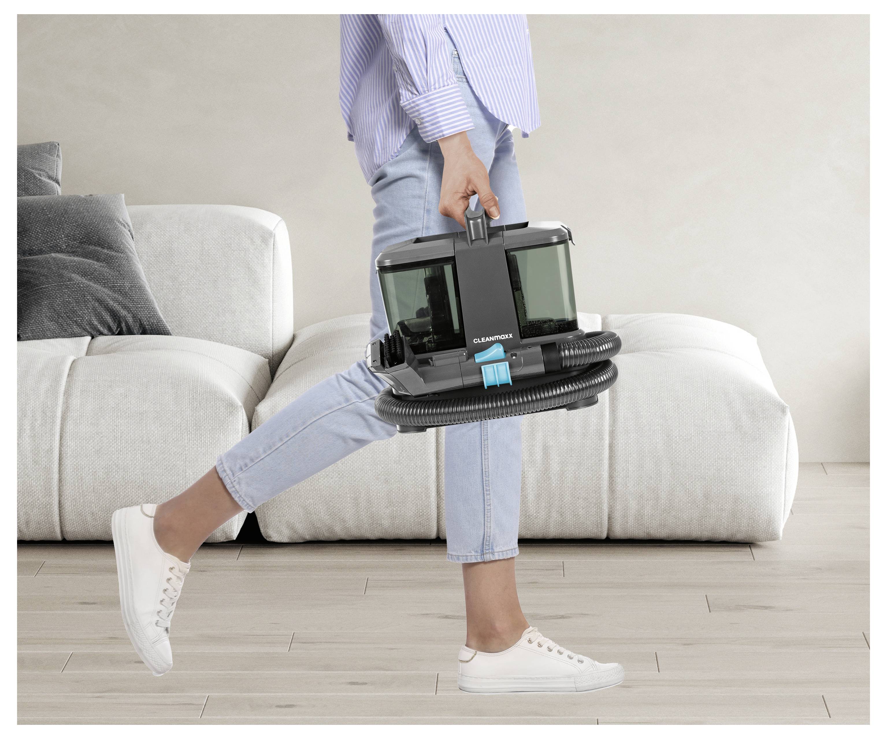 Eine Person in lässiger Kleidung geht im Innenraum und trägt einen kompakten, modernen Staubsauger neben einem weißen Sofa in einem Wohnzimmer.