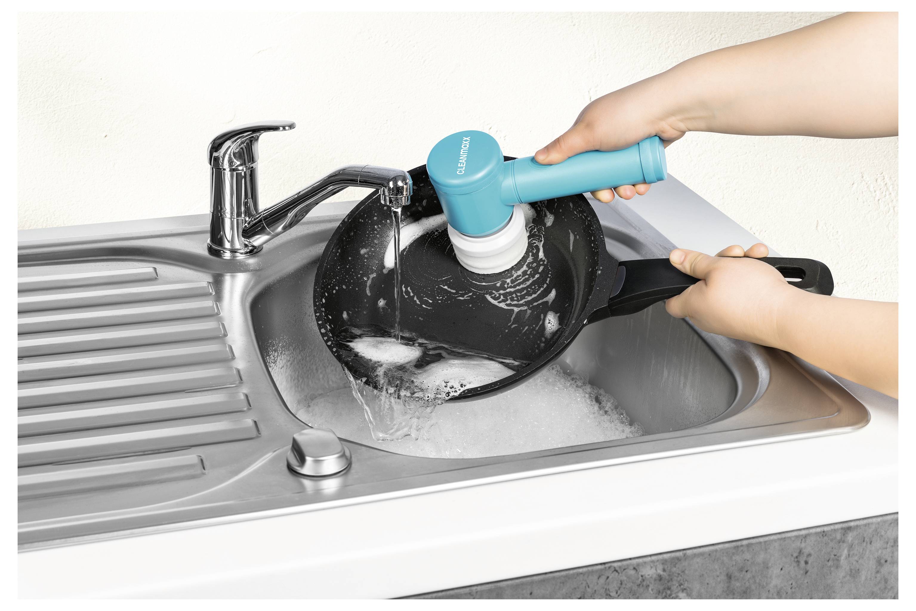 Hände benutzen eine blaue elektrische Reinigungsbürste, um eine Bratpfanne mit Seife unter einem laufenden Wasserhahn in einer silbernen Spüle zu reinigen.