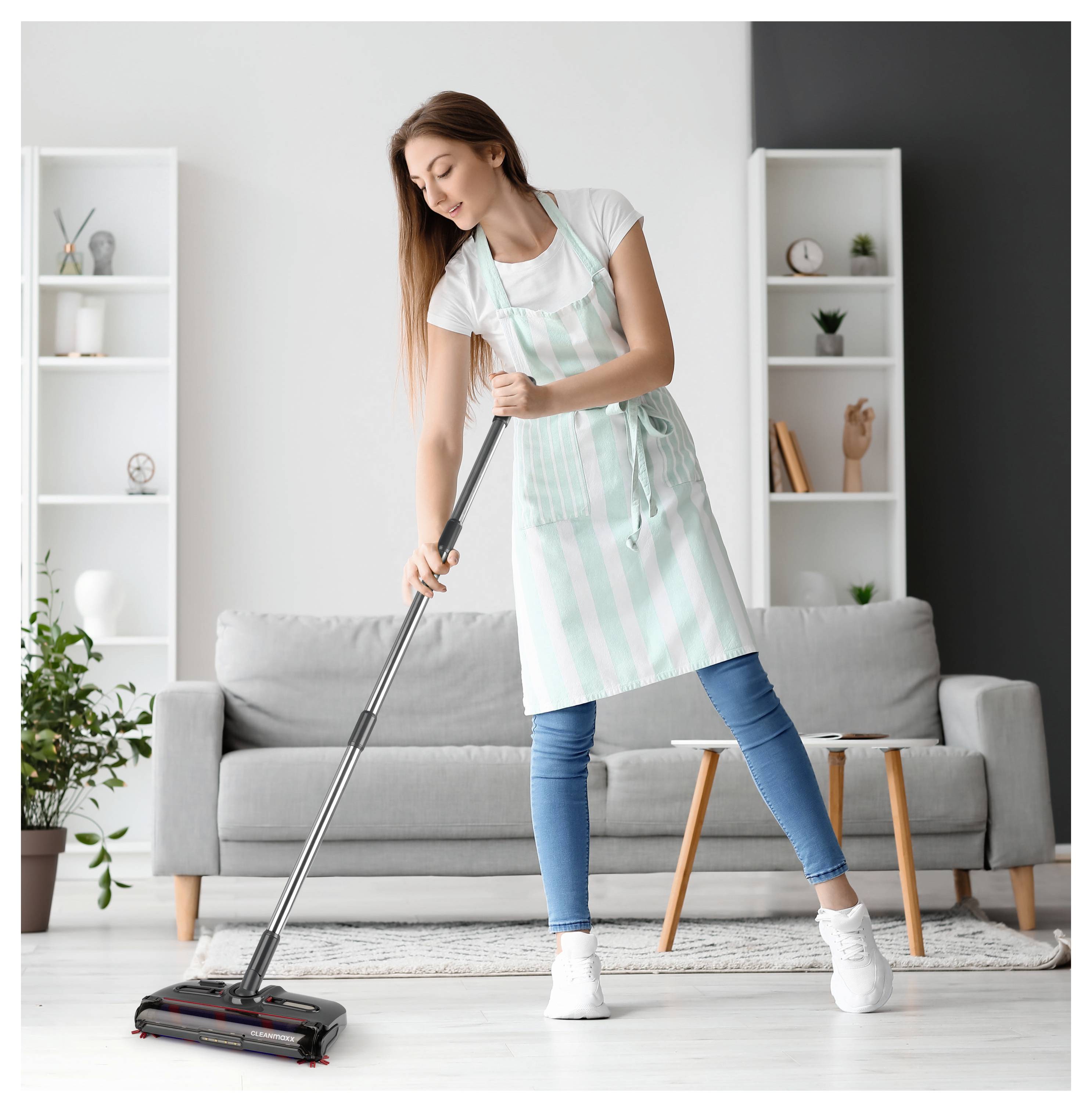 Eine Frau in einer gestreiften Schürze staubsaugt ein Wohnzimmer mit moderner Einrichtung, darunter ein graues Sofa, Pflanzen und ein Bücherregal.