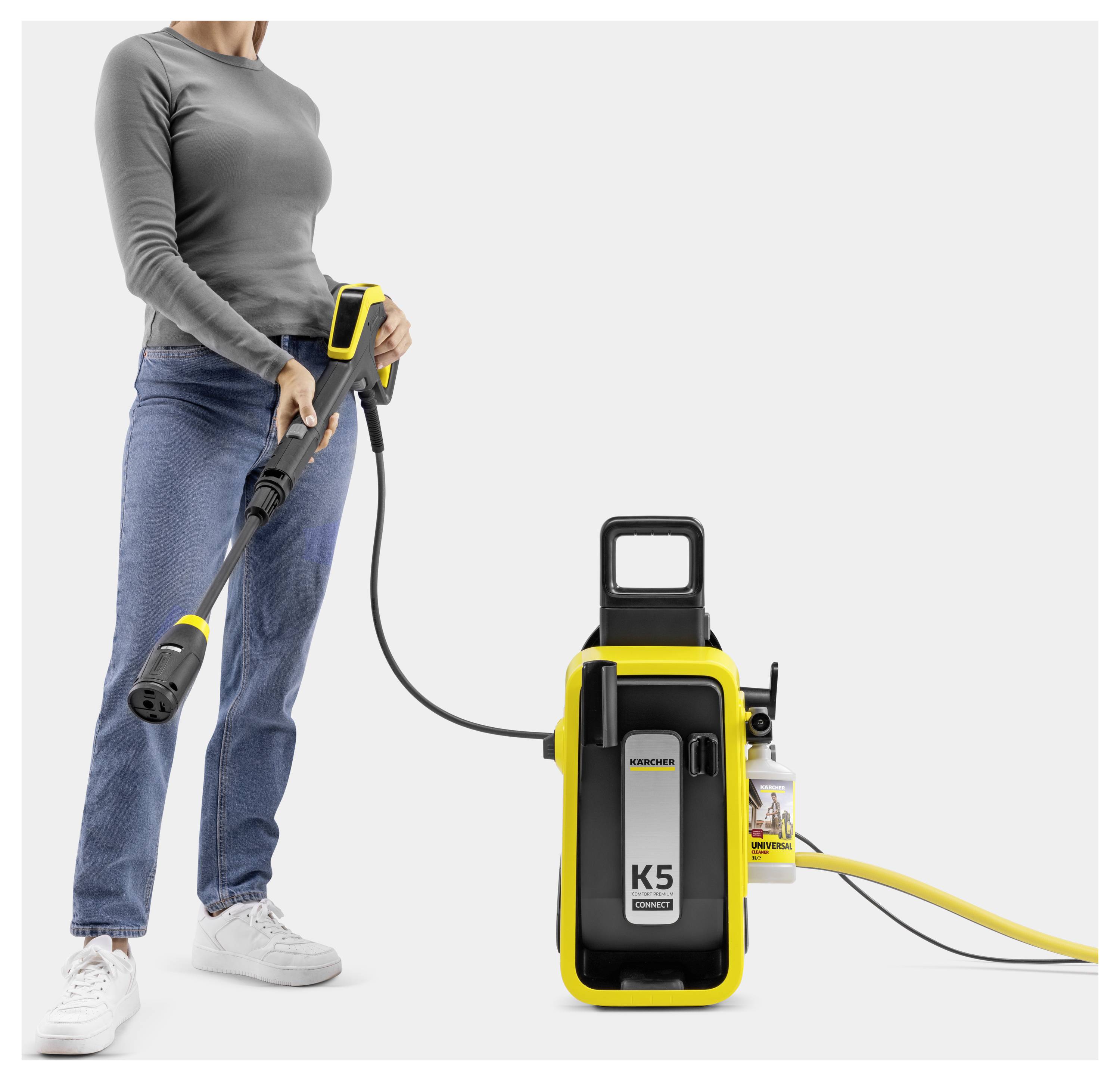 Eine Person, die einen Kärcher K5 Hochdruckreiniger nutzt, der durch einen Schlauch angeschlossen ist, hält die Auslösepistole, gekleidet in Freizeitkleidung, vor einem neutralen Hintergrund.