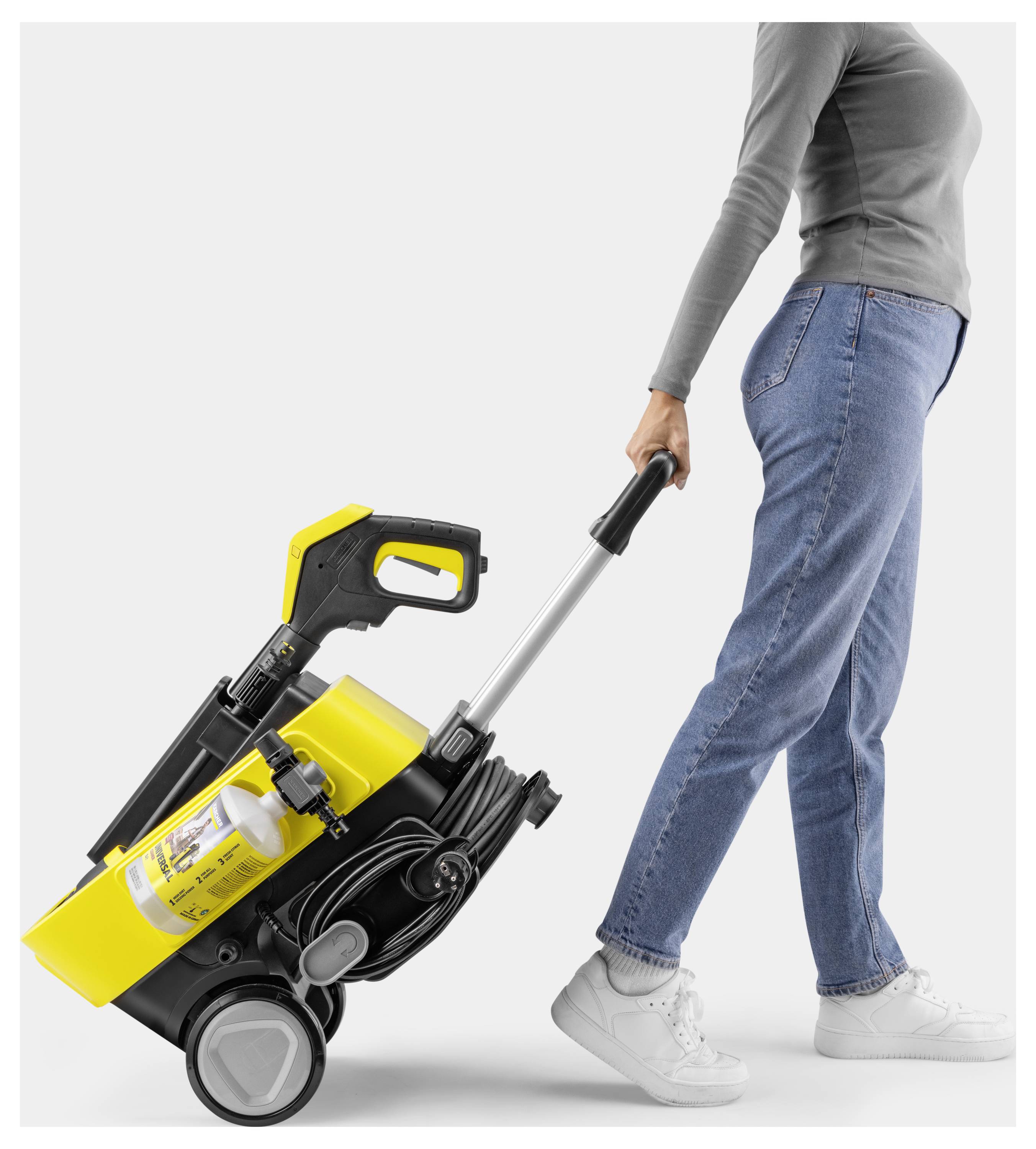 Eine Person in Jeans und grauem Oberteil zieht einen gelben Staubsauger auf Rädern mit metallischem Griff, was seine Tragbarkeit unterstreicht.