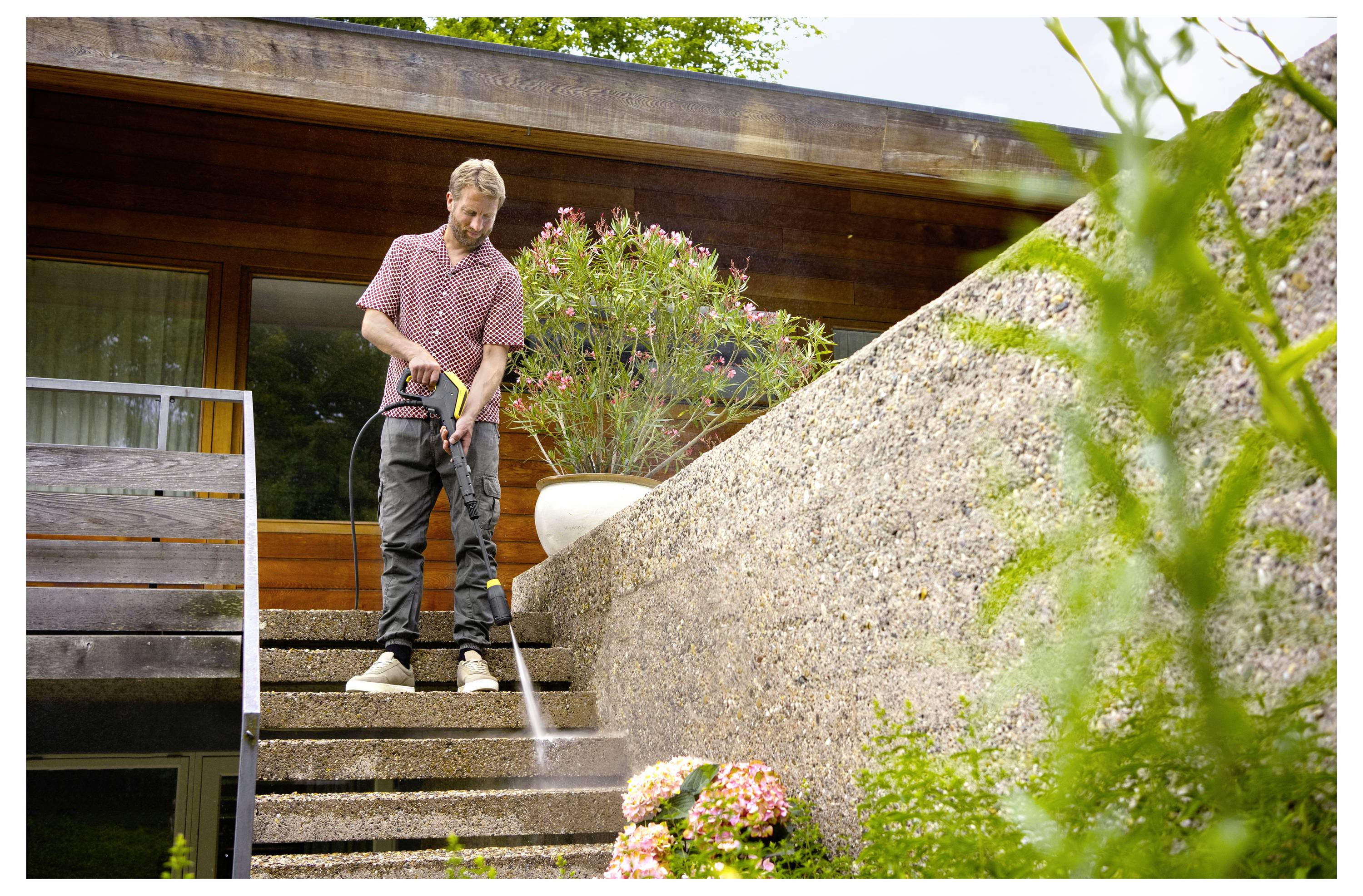 Eine Person verwendet einen Hochdruckreiniger, um Betonstufen vor einem modernen Holzhaus zu reinigen, umgeben von Pflanzen und Blumen.