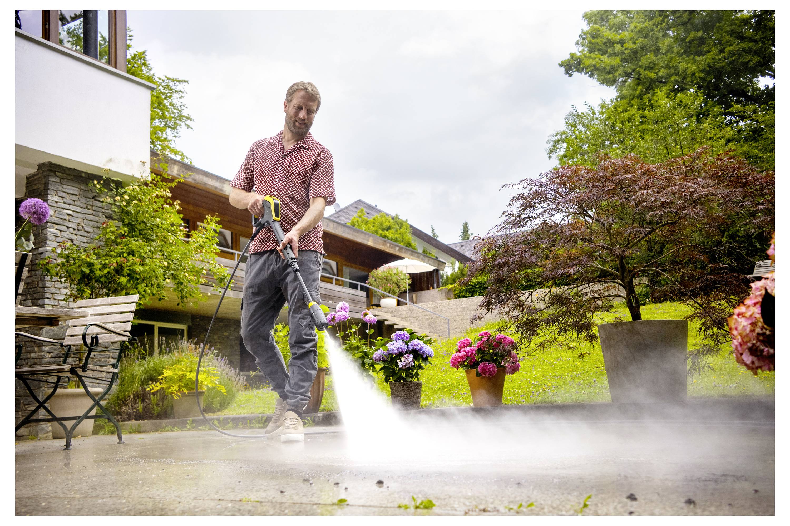 Eine Person in einem Garten, die einen Hochdruckreiniger verwendet, um eine Terrasse zu reinigen. Blumen und Bäume umgeben das Gebiet und schaffen eine ruhige Atmosphäre.