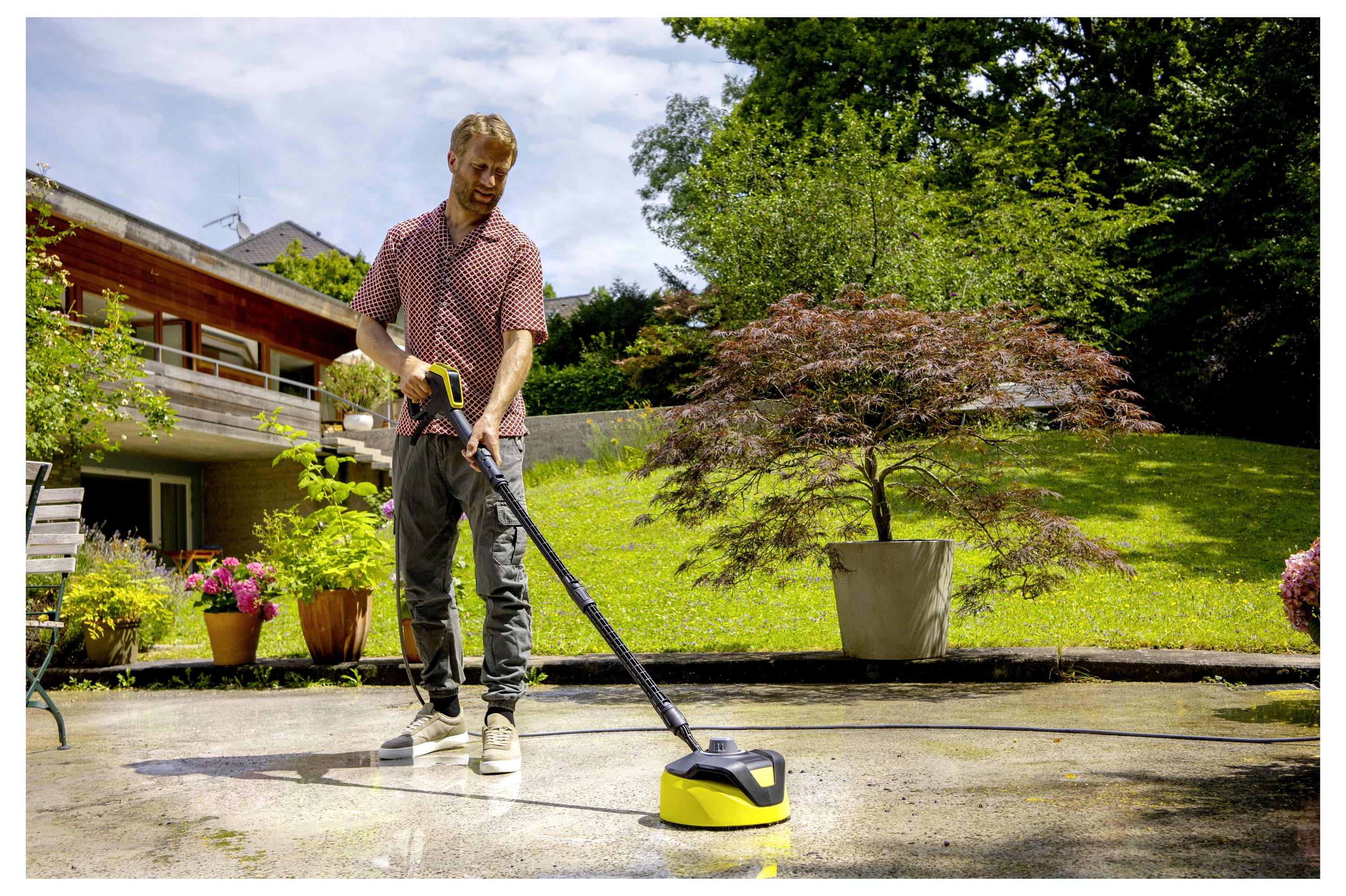 Eine Person benutzt einen Hochdruckreiniger, um eine Terrasse in einem Garten mit Topfpflanzen und einem großen Baum im Hintergrund zu reinigen.