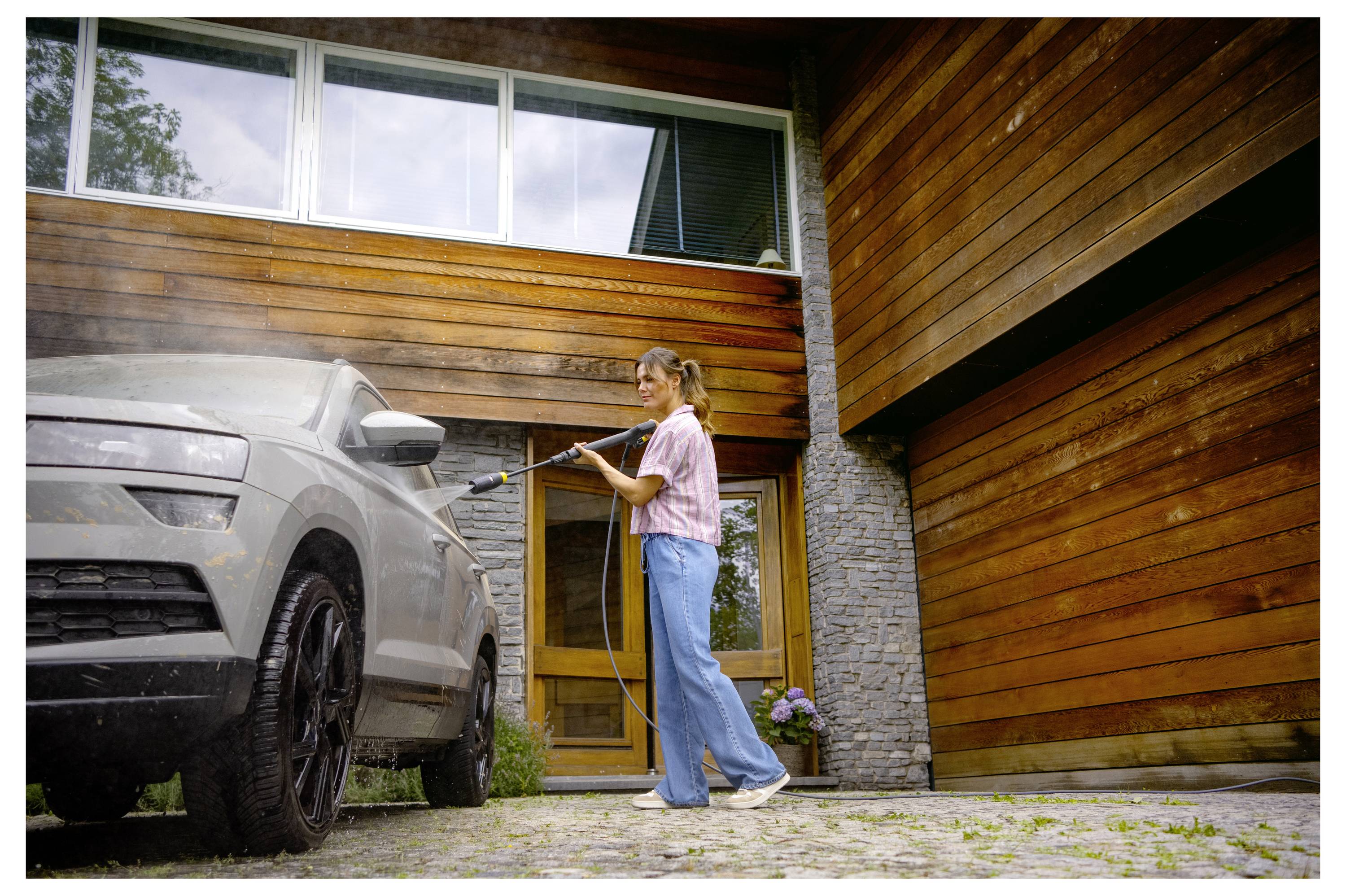 Eine Person reinigt ein Auto mit einem Hochdruckreiniger vor einem modernen Holzhaus, wobei große Fenster im Hintergrund sichtbar sind.