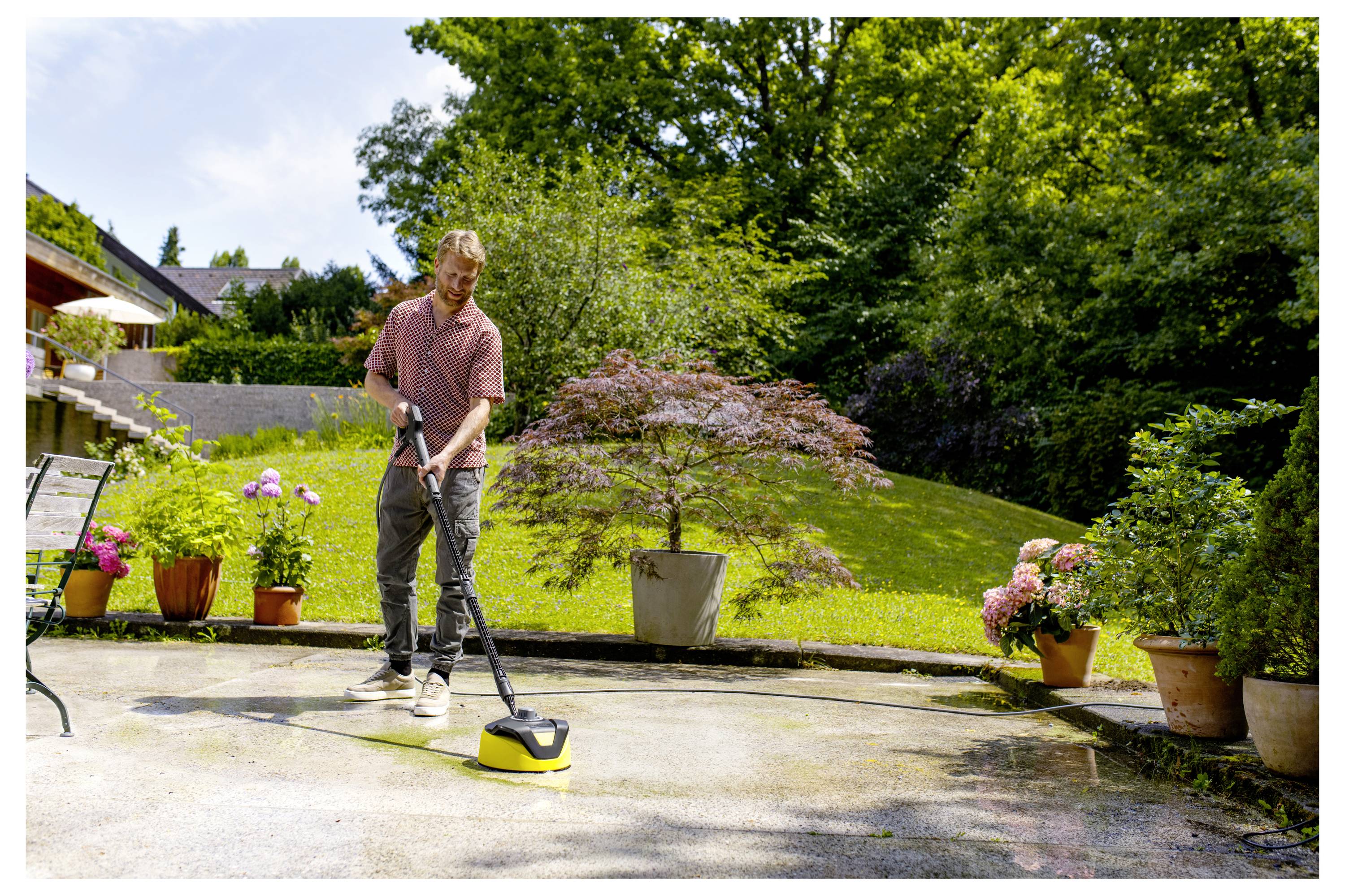 Ein Mann benutzt einen Hochdruckreiniger, um eine Terrasse in einem sonnigen Garten mit Bäumen und Topfpflanzen herum zu reinigen.