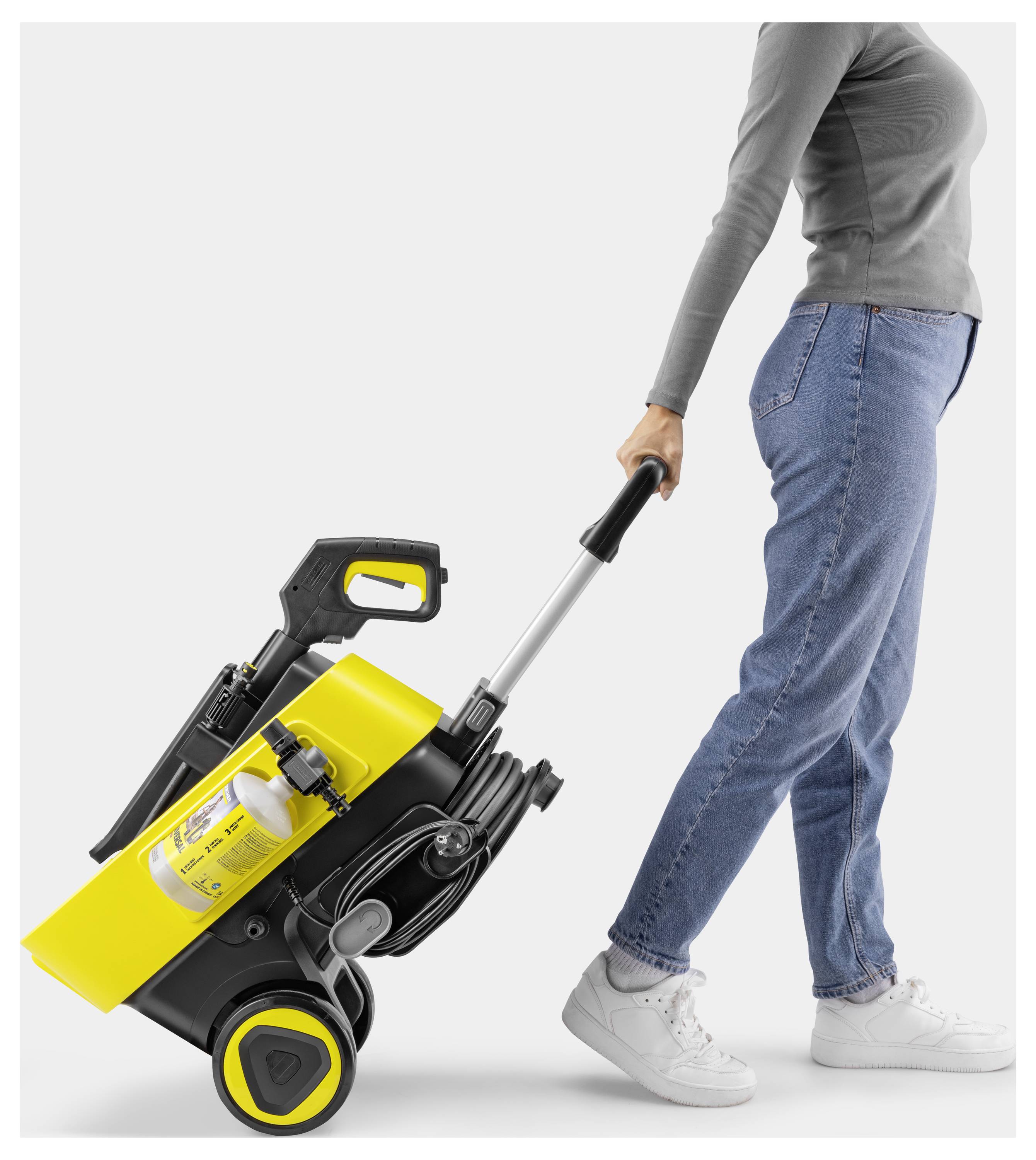 Eine Person in Jeans und grauem Langarmshirt zieht eine gelb-schwarze tragbare Reinigungsmaschine mit großen Rädern auf weißem Hintergrund.