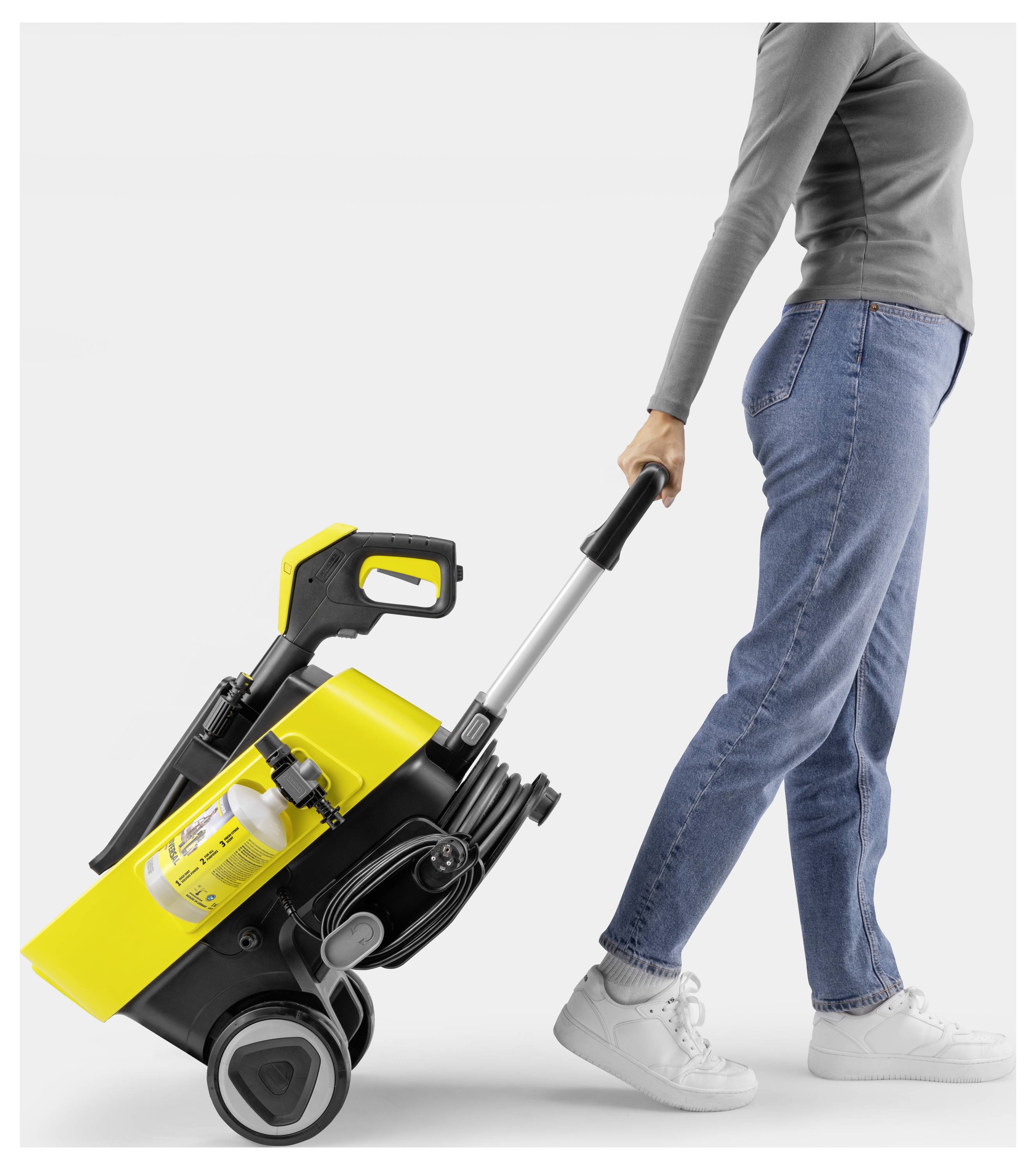Eine Person zieht einen gelb-schwarz gestreiften fahrbaren Hochdruckreiniger und trägt dabei blaue Jeans, weiße Sneaker und ein graues Langarmshirt.