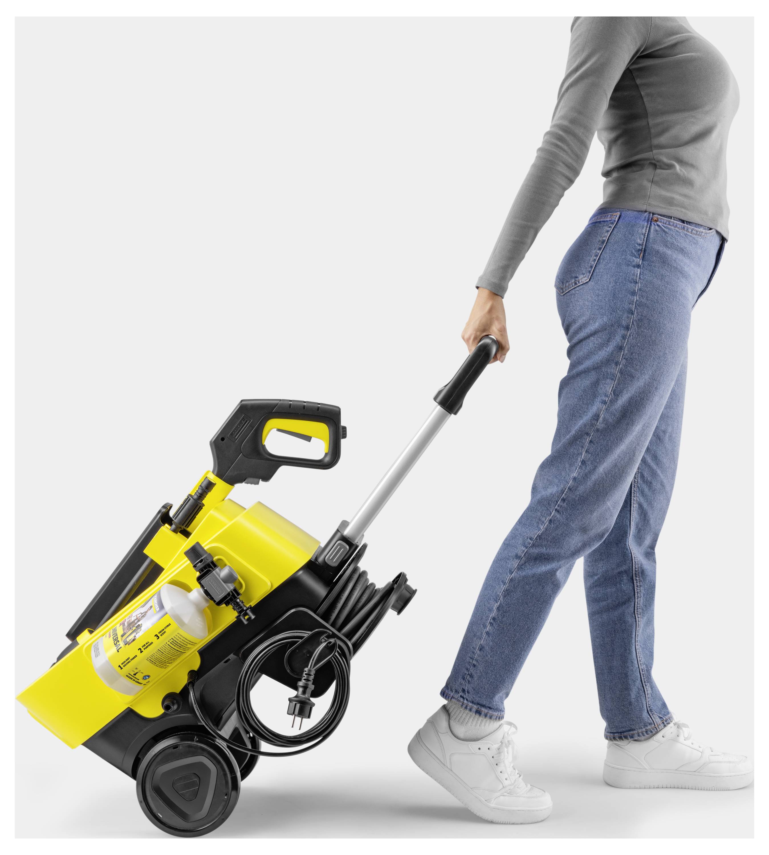 Eine Person in Jeans und grauem Oberteil zieht einen gelb-schwarzen tragbaren Hochdruckreiniger auf Rädern vor einem einfachen Hintergrund.