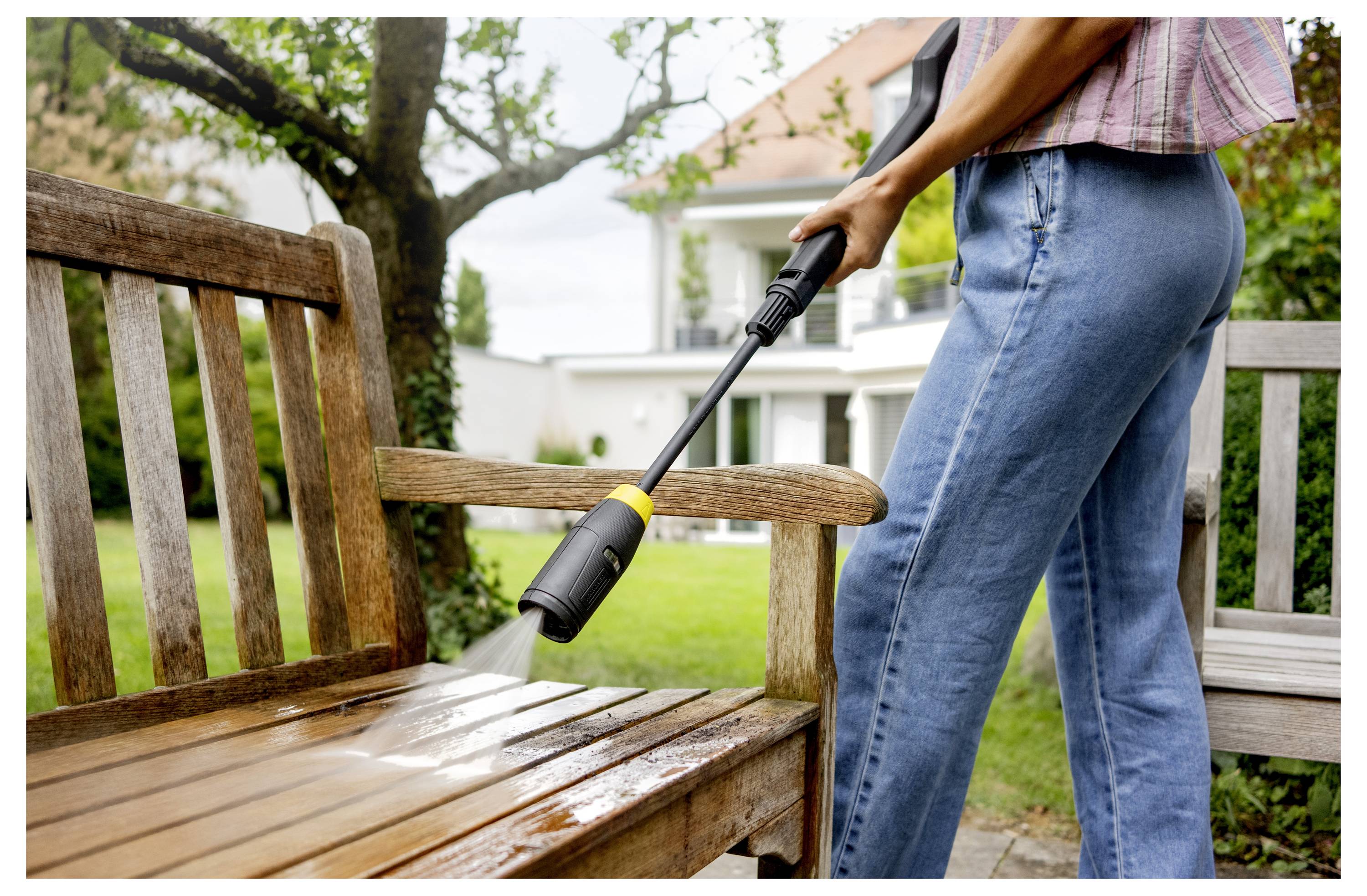 Eine Person benutzt einen Hochdruckreiniger, um eine Holzbank in einem Garten mit Bäumen und einem Haus im Hintergrund zu reinigen.