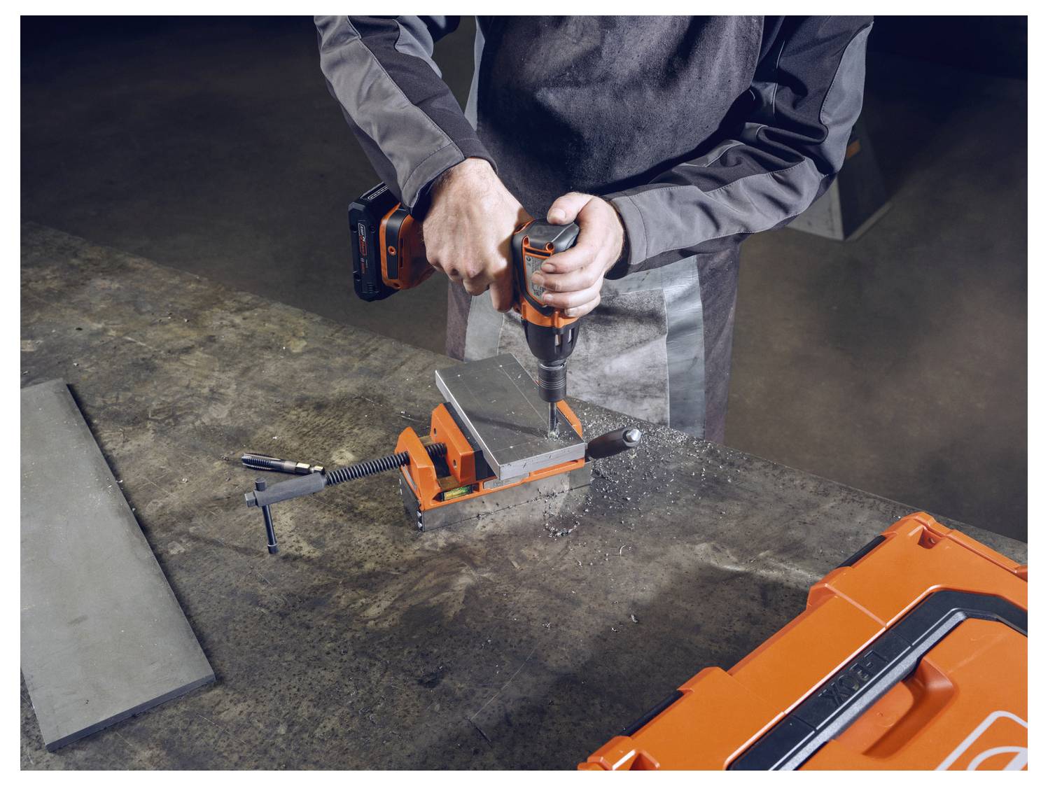 Eine Person bedient eine orange elektrische Bohrmaschine auf einem Metalltisch, wobei ein Metallteil eingespannt ist, umgeben von einer orange Werkzeugbox.