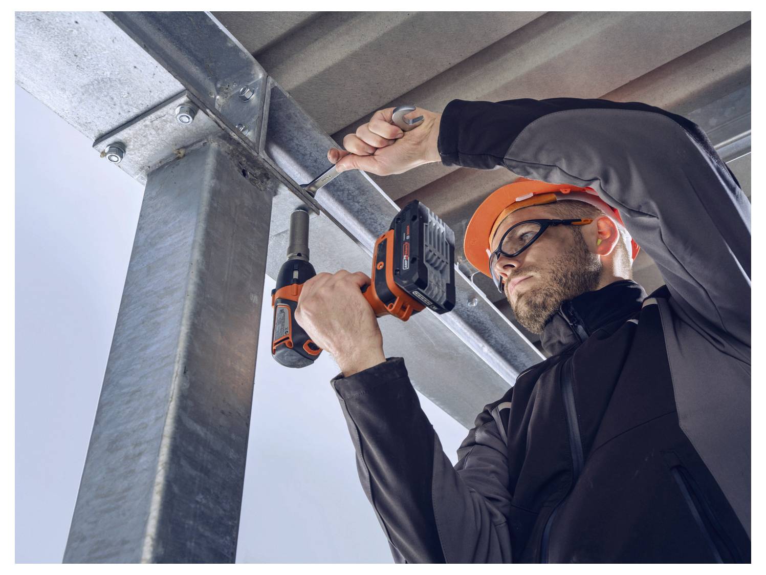 Eine Person, die Schutzausrüstung trägt, benutzt eine Elektrobohrmaschine, um einen Metallträger in einer industriellen Umgebung zu befestigen.