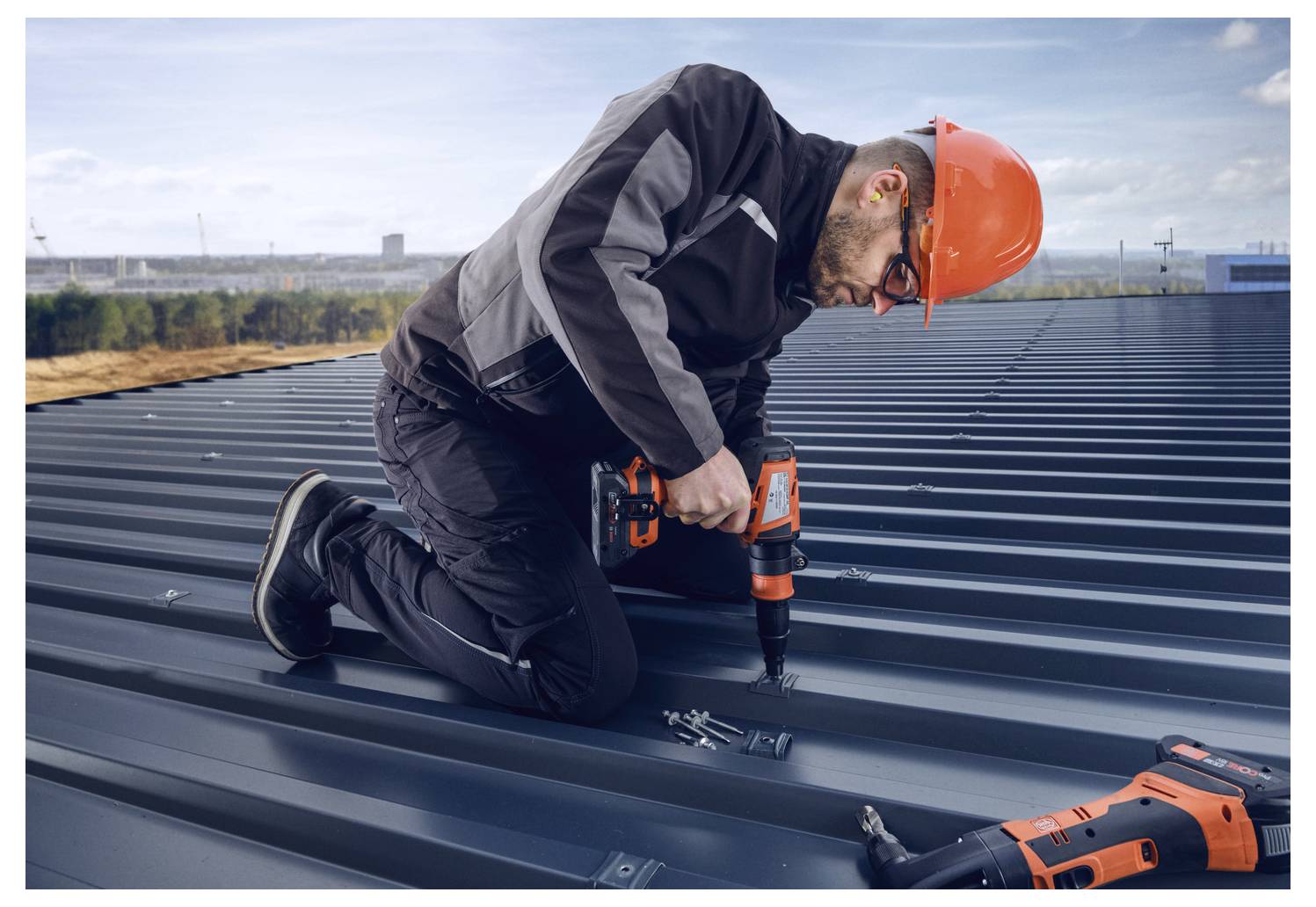 Ein Bauarbeiter in einem Schutzhelm benutzt einen Elektroschrauber auf einem Metalldach.
