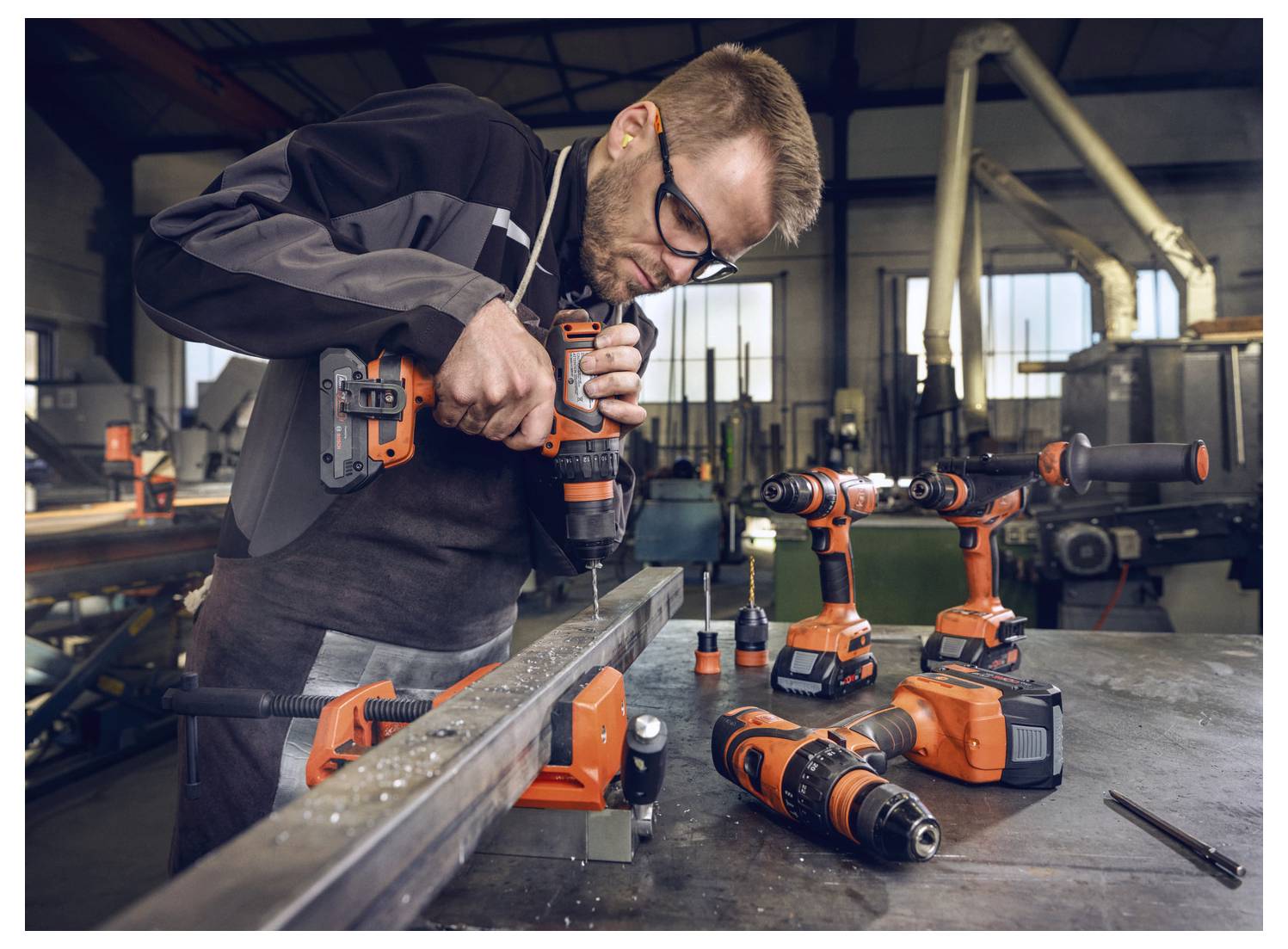 Ein Arbeiter in Schutzbrille bohrt mit einem Bohrer an einem Metallträger in einer Werkstatt. Andere Bohrer und Werkzeuge sind auf der Werkbank sichtbar.