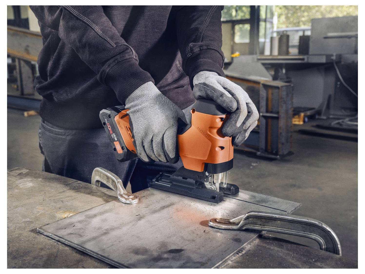 Eine Person mit Handschuhen benutzt ein elektrisches Werkzeug, um ein Metallblech in einer Werkstatt zu schneiden, umgeben von Industrieausrüstung.