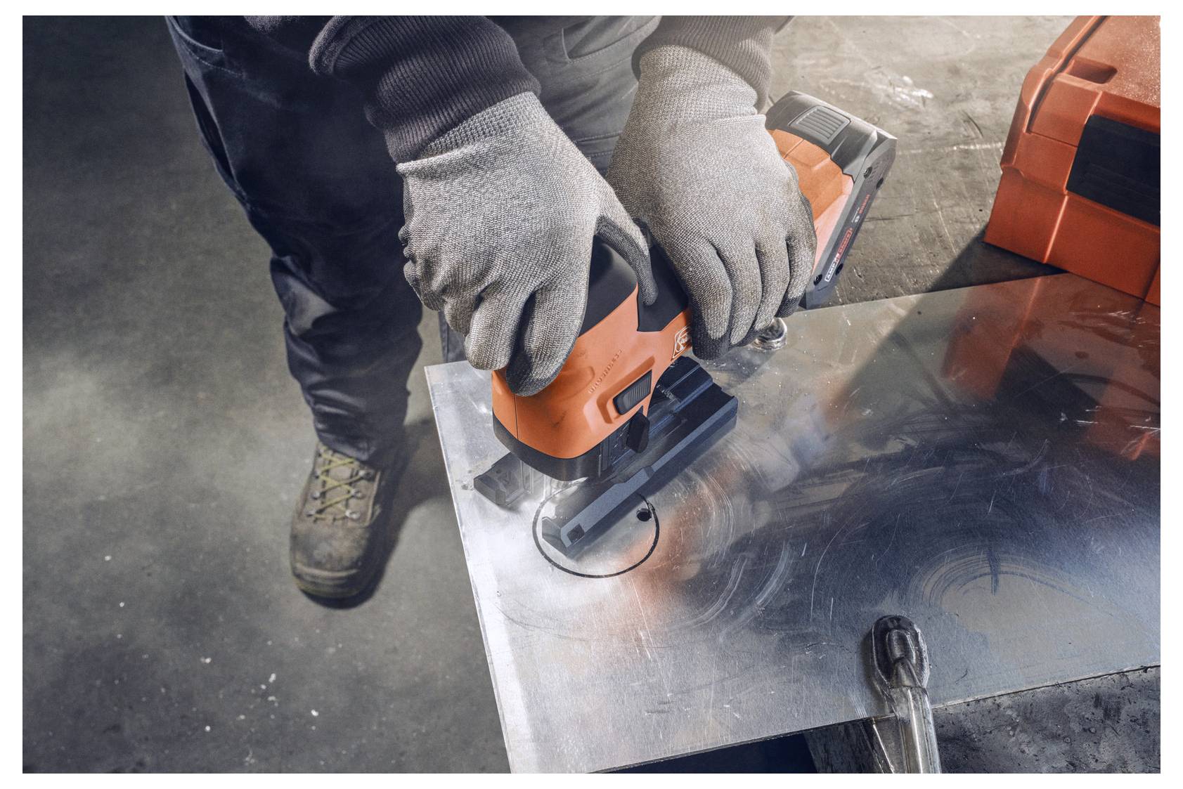 Eine Person, die Handschuhe trägt, benutzt eine orangefarbene elektrische Stichsäge, um eine kreisförmige Form in ein Metallblech auf einer Werkbank zu schneiden.