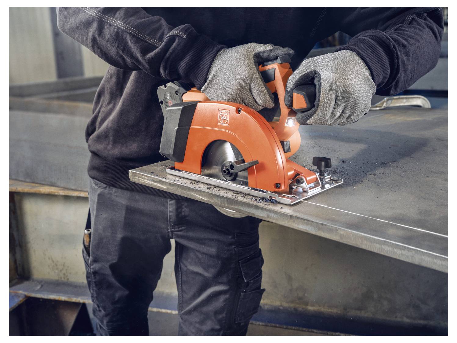 Eine Person mit Handschuhen benutzt eine orangefarbene Kreissäge, um ein Metallblech in einer industriellen Umgebung zu schneiden.