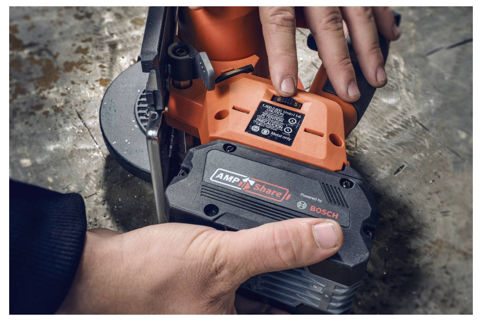 Eine Person drückt einen Knopf an einem Elektrowerkzeug mit dem Bosch-Logo. Eine andere Hand hält das Werkzeug auf einer rauen Oberfläche fest.