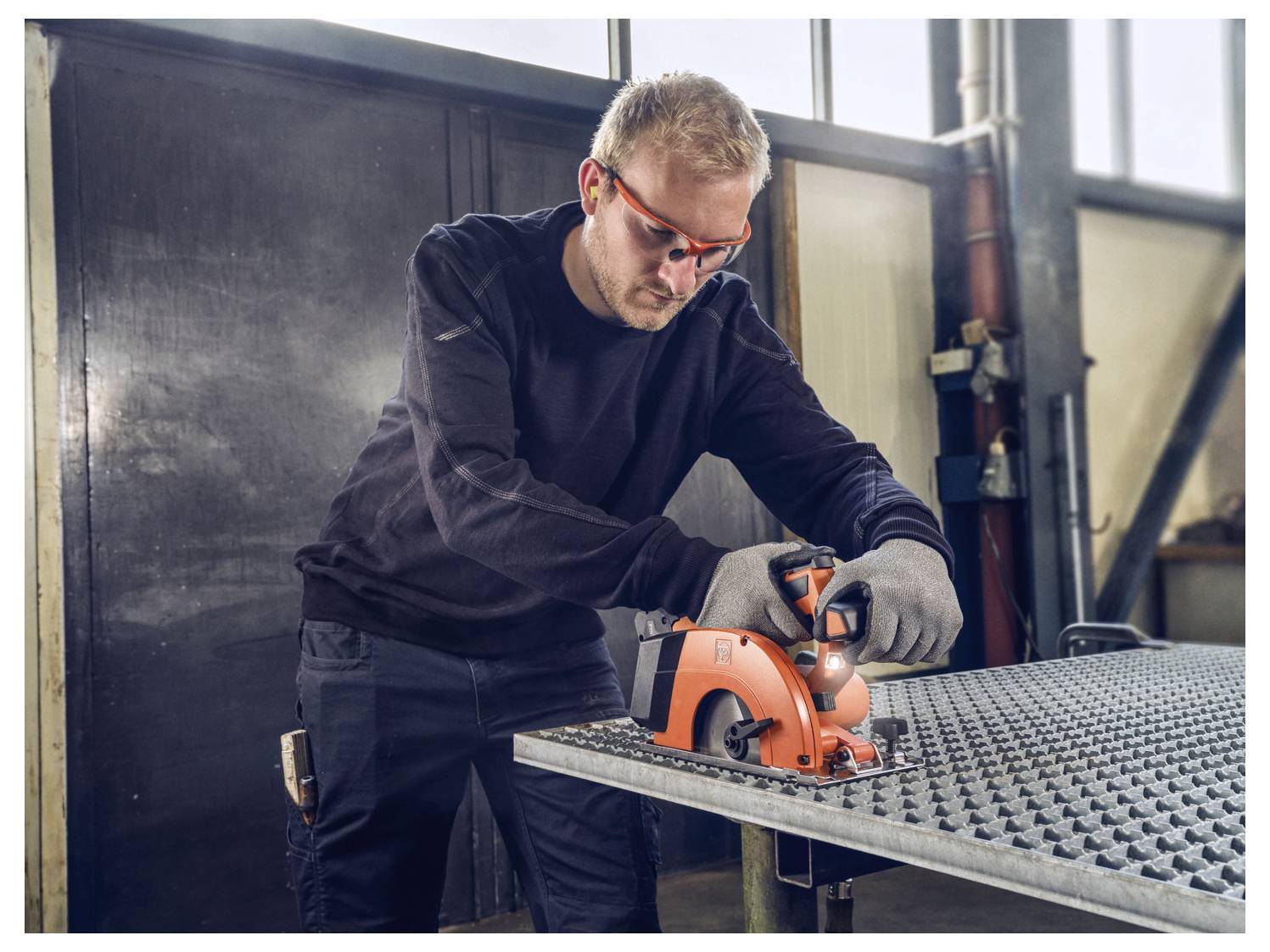 Ein Mann in Schutzausrüstung verwendet ein Elektrowerkzeug auf einer Metalloberfläche in einer industriellen Umgebung.