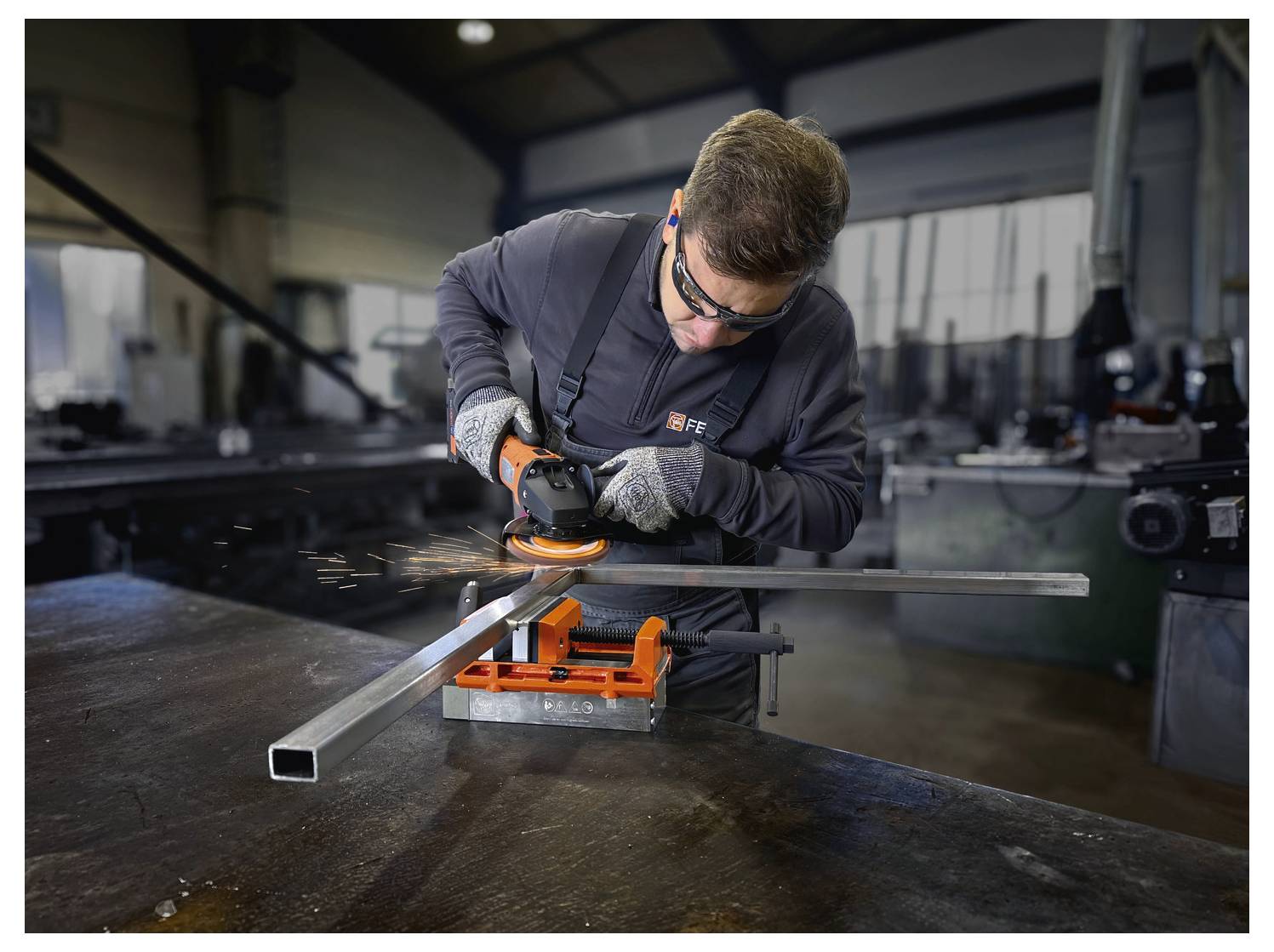 Eine Person in einer Werkstatt trägt Schutzausrüstung und nutzt einen Winkelschleiffer, um ein Metallquadratrohr zu schneiden, das auf einem Tisch befestigt ist. Funken sind sichtbar.