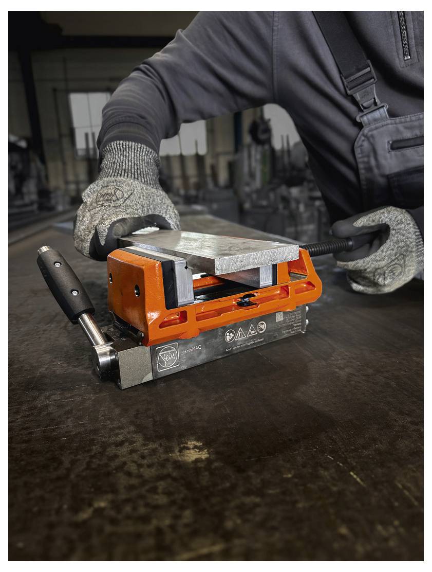 Ein Arbeiter benutzt ein orangefarbenes Metallschleifwerkzeug auf einem großen Tisch, trägt Handschuhe und eine dunkle Schürze in einer industriellen Umgebung.