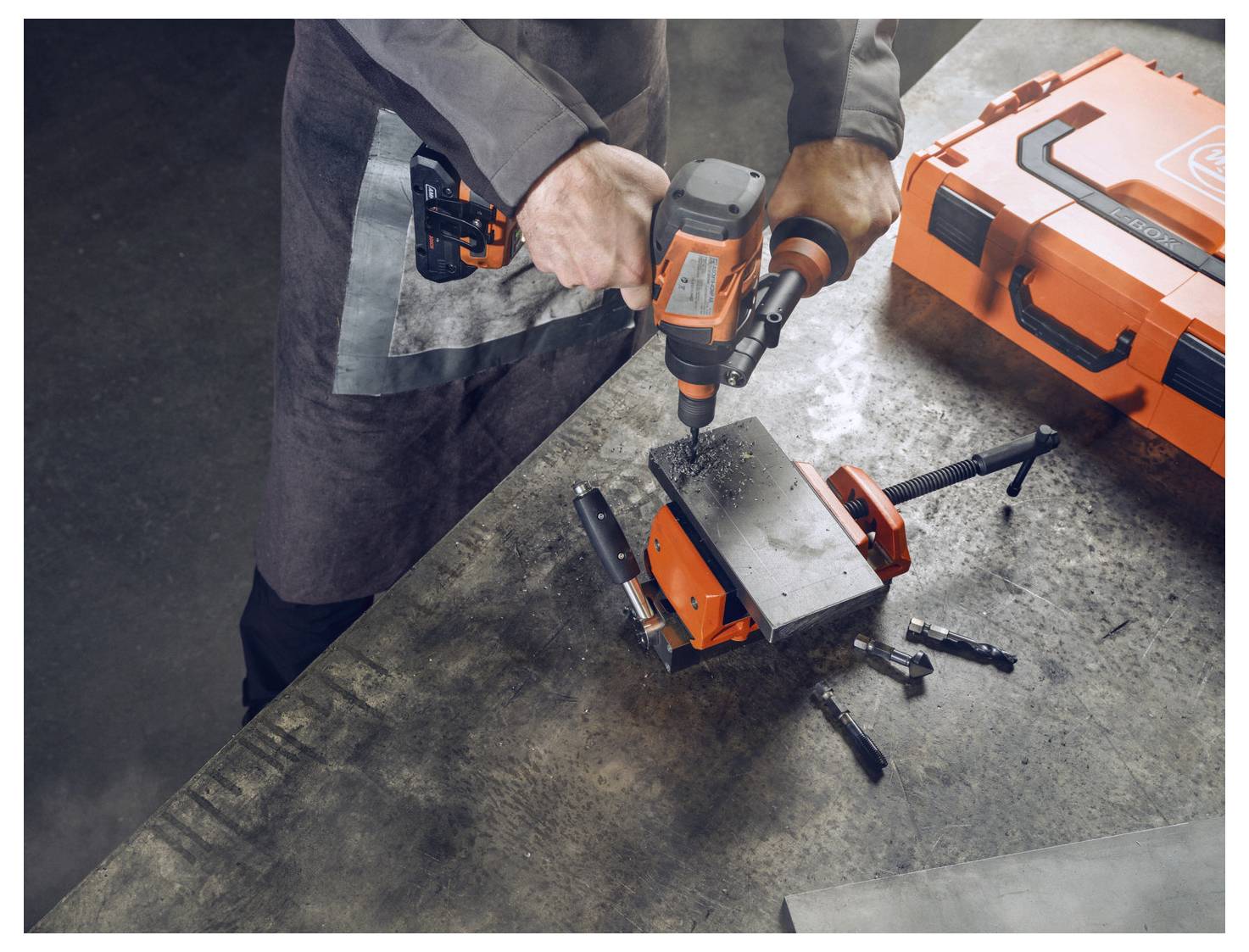 Eine Person benutzt einen orangefarbenen Elektroschrauber an Metall, das in einer Werkbank eingespannt ist, mit Werkzeugen und einem orangefarbenen Koffer in der Nähe.
