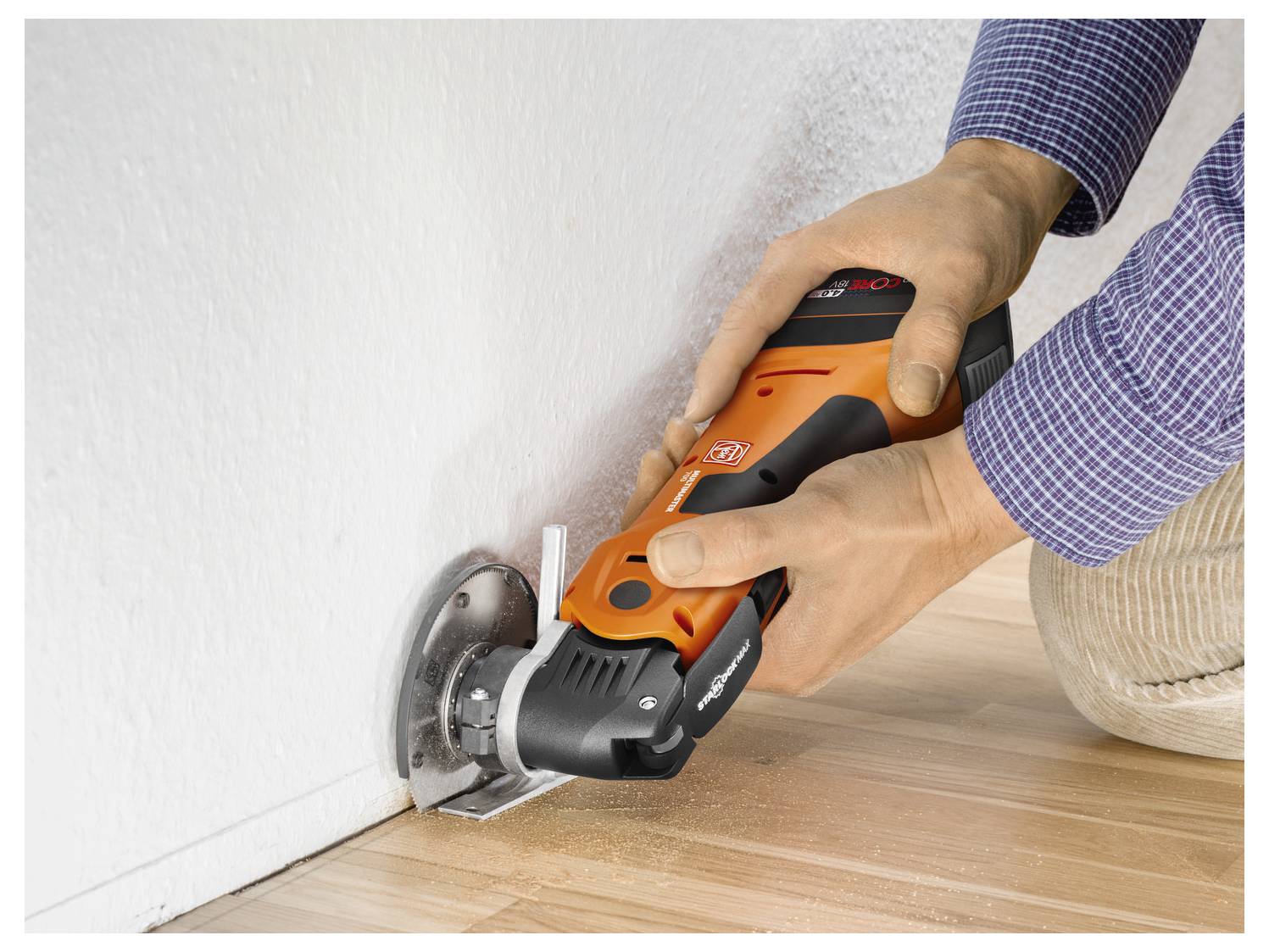 Eine Person benutzt ein orangefarbenes Elektrowerkzeug, um entlang einer weißen Wand in einen Holzboden zu schneiden.